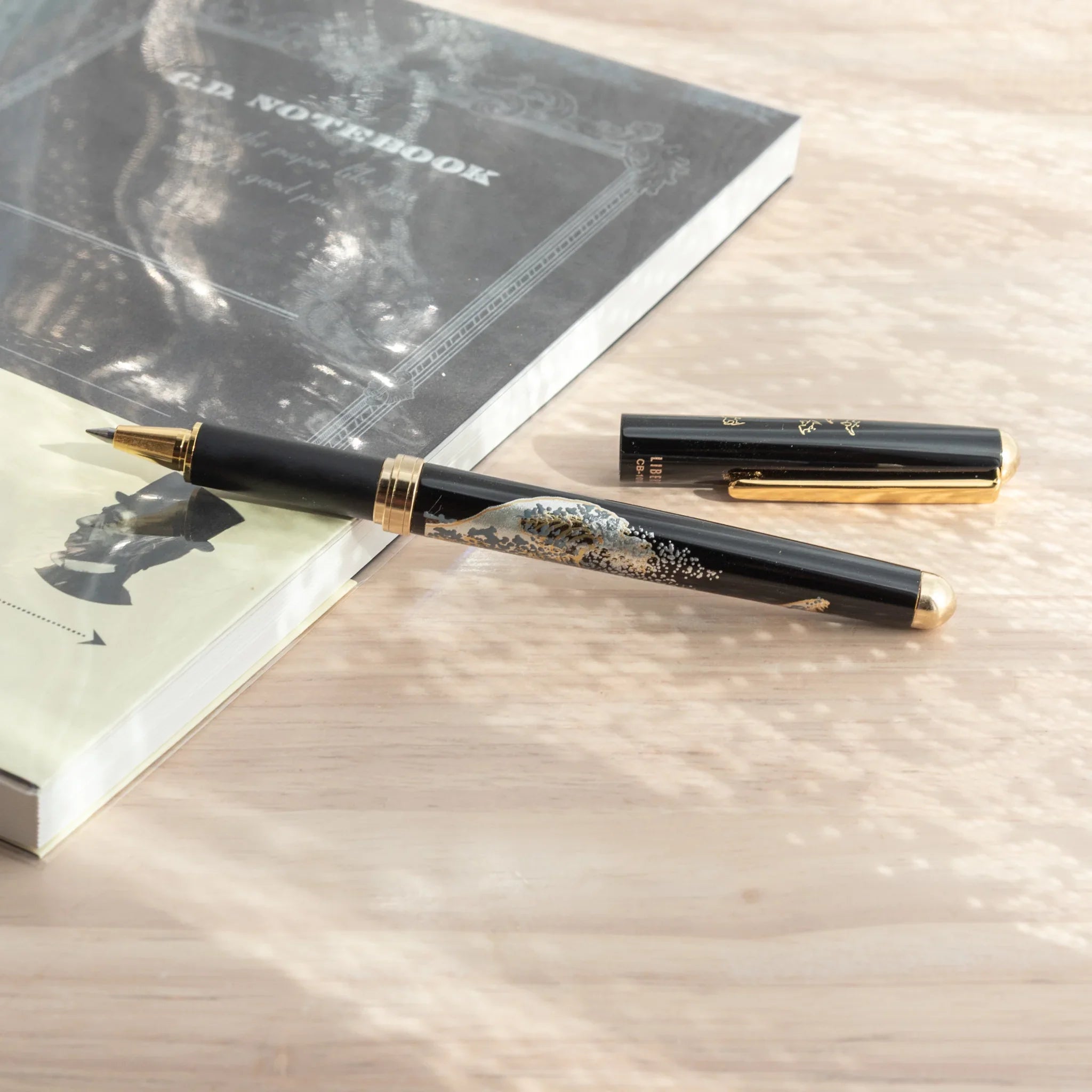 Maeju Yamanaka ballpoint pen body with Great Wave silkscreen, cap separated, on wood surface beside dark G.D. Notebook