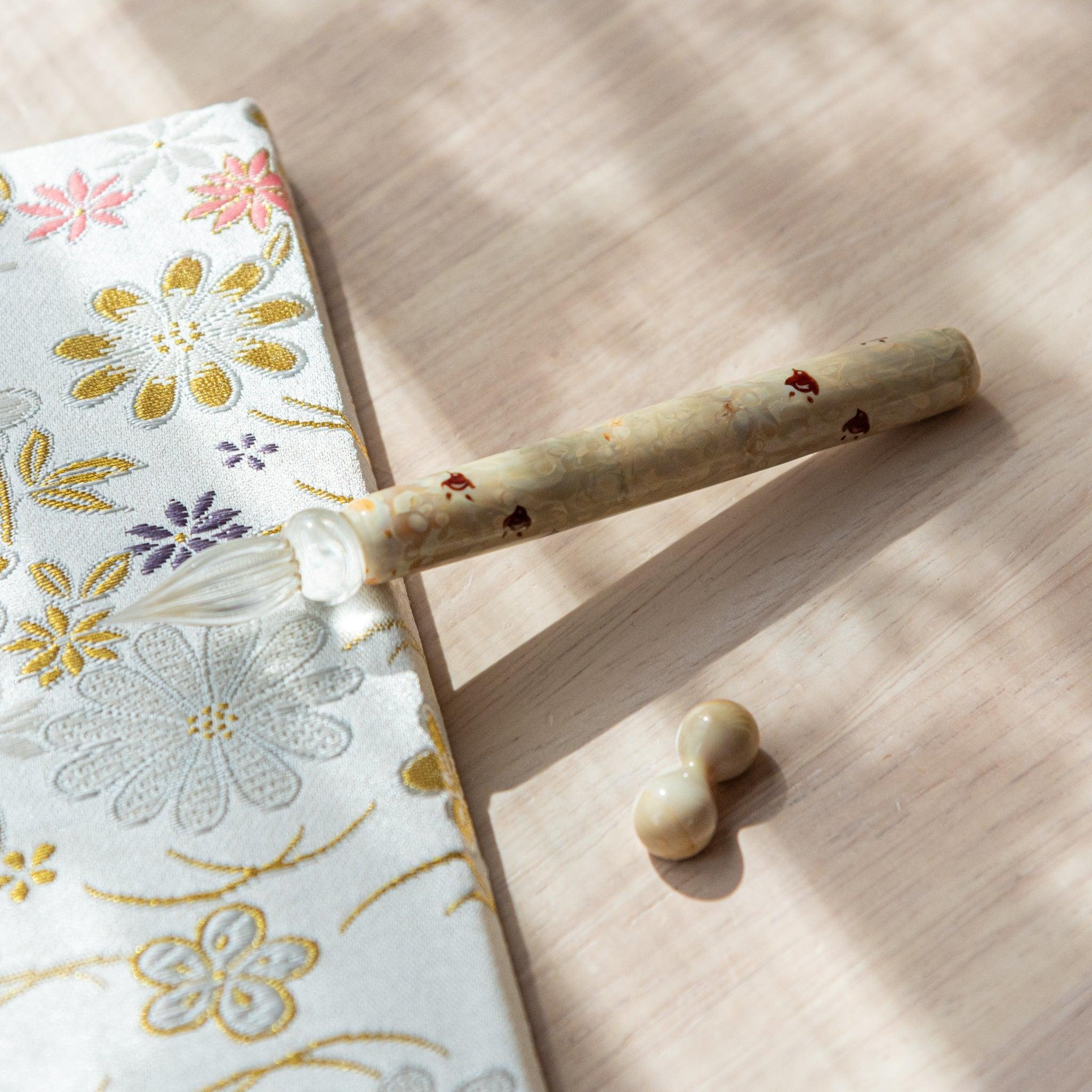 Flatlay of ivory chidori glass dip pen displayed on decorative embroidered fabric with pen rest components visible