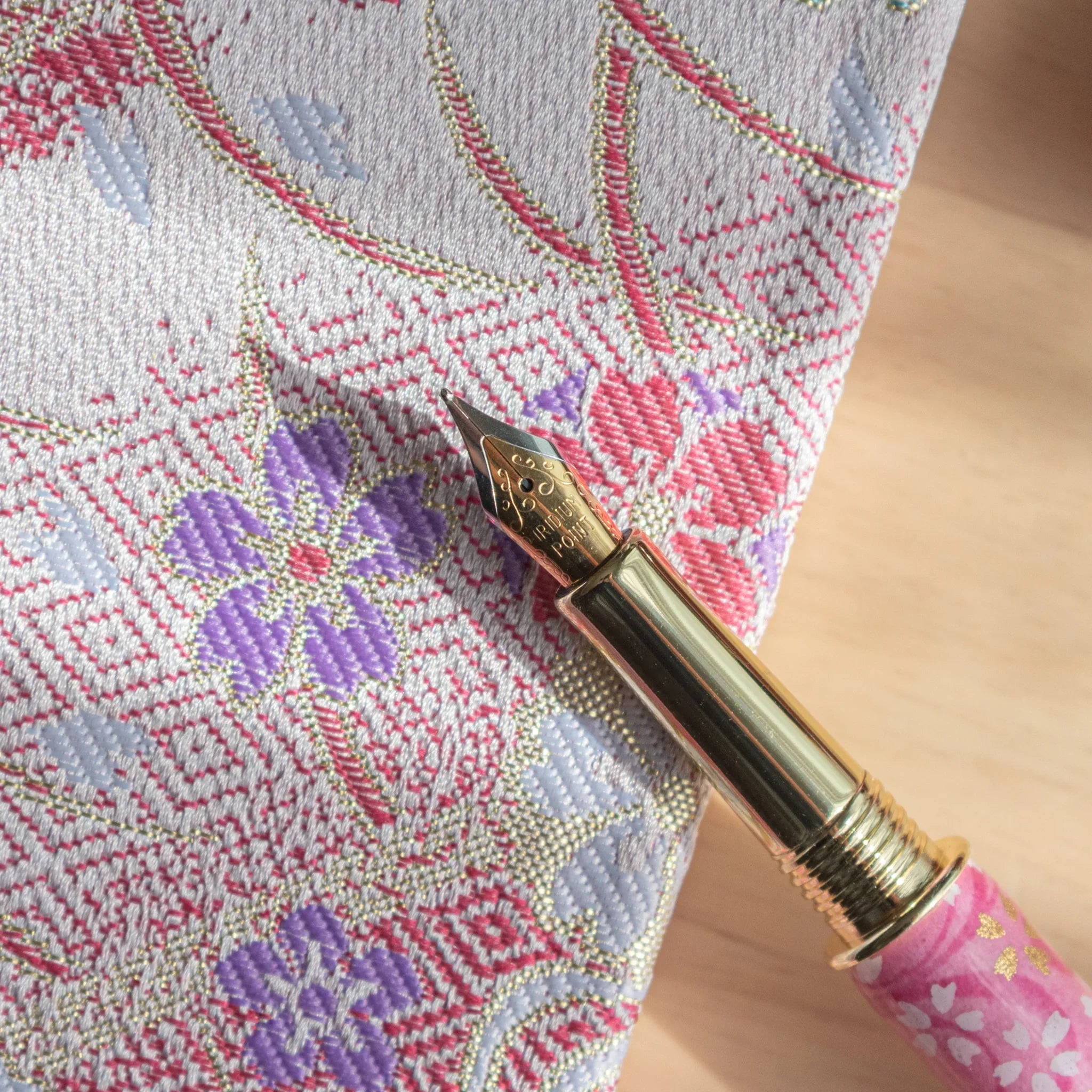 Close-up of the gold nib with engraved text and pink Mino washi barrel of a Japanese fountain pen on a woven textile