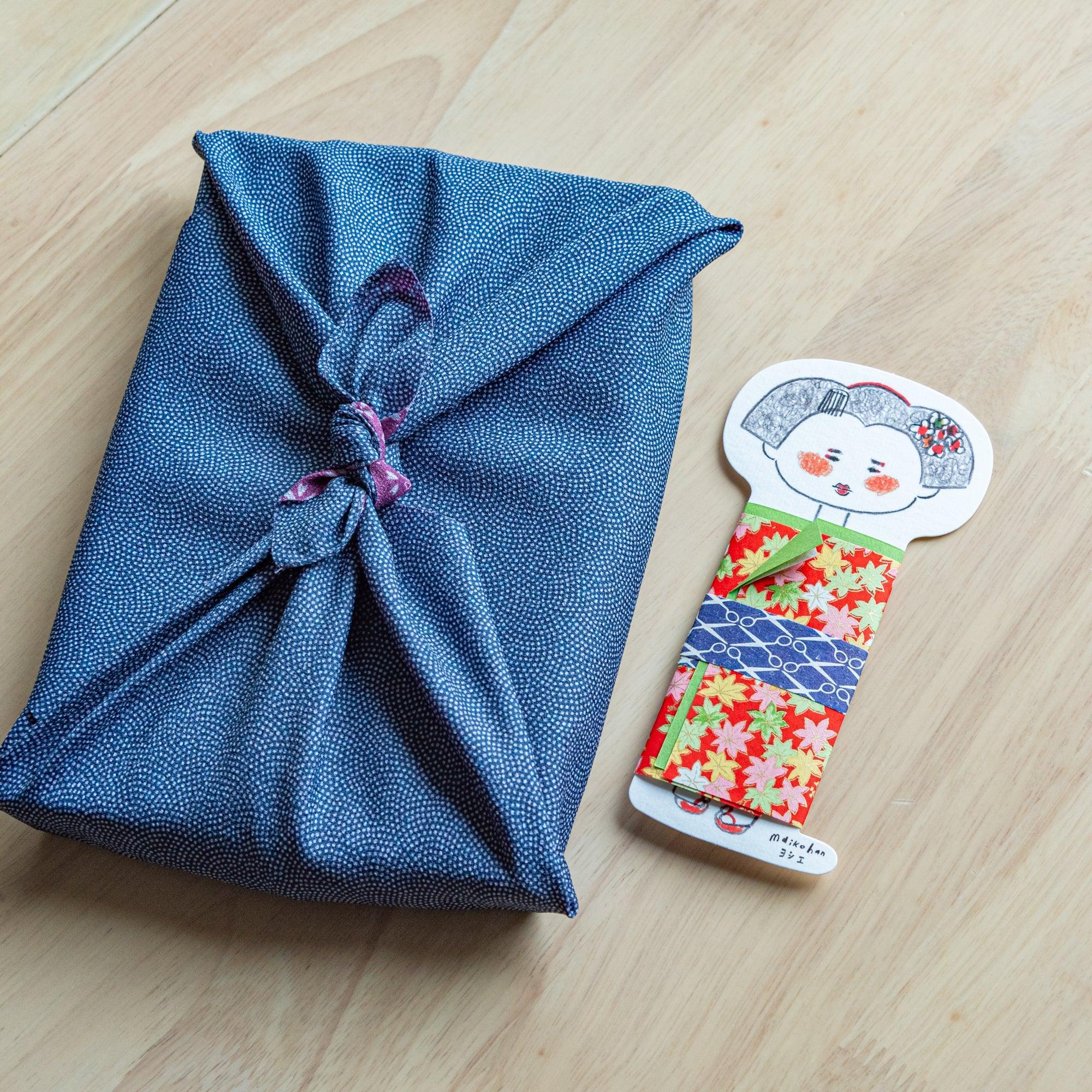 Maiko die-cut message card with red maple leaf yuzen washi kimono standing beside a furoshiki-wrapped gift on a wood surface