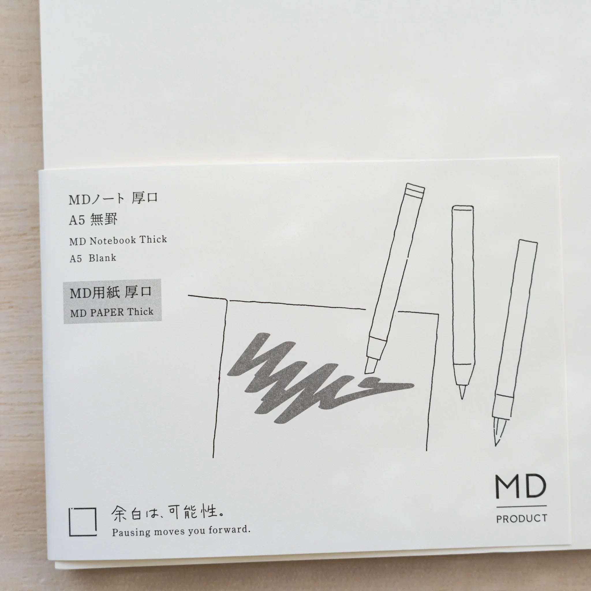 Close-up of Midori MD Notebook Thick paper belly band, MD PAPER Thick text and line drawing of three pens writing