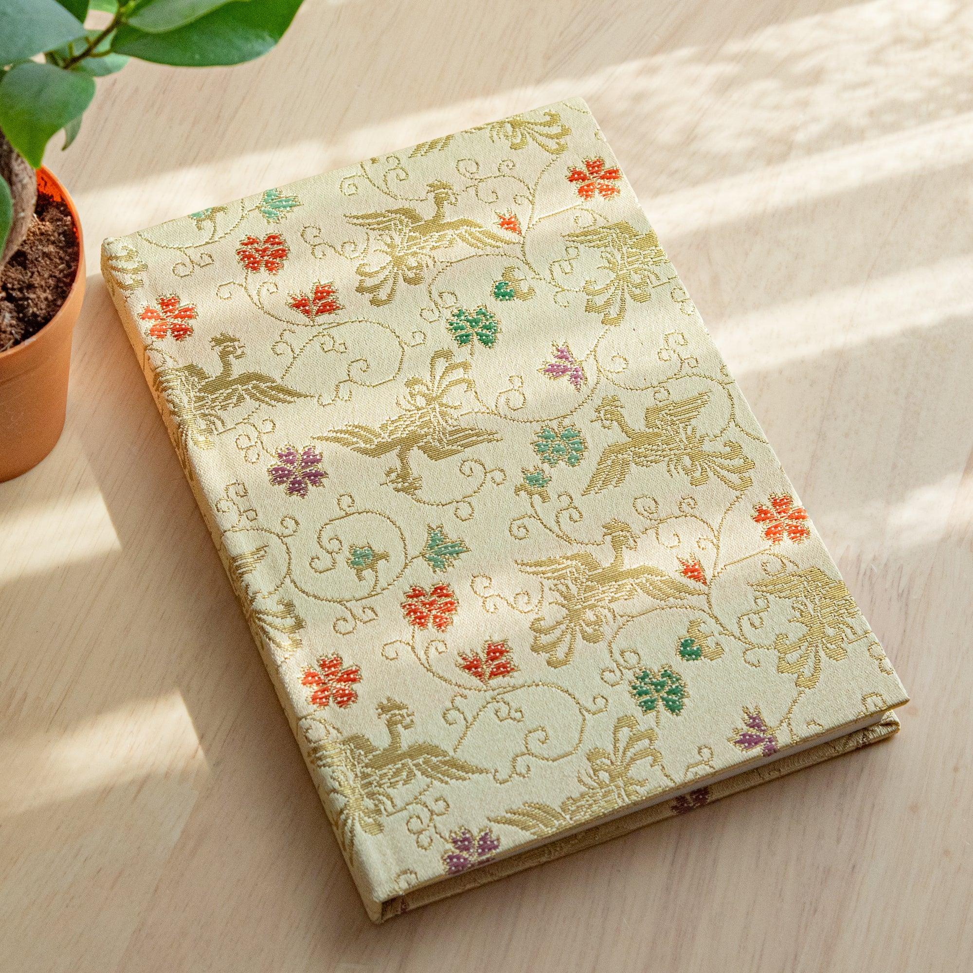 Japanese notebook with pale gold Nishijin woven cover showing phoenix and arabesque scrollwork, angled on wood with plant