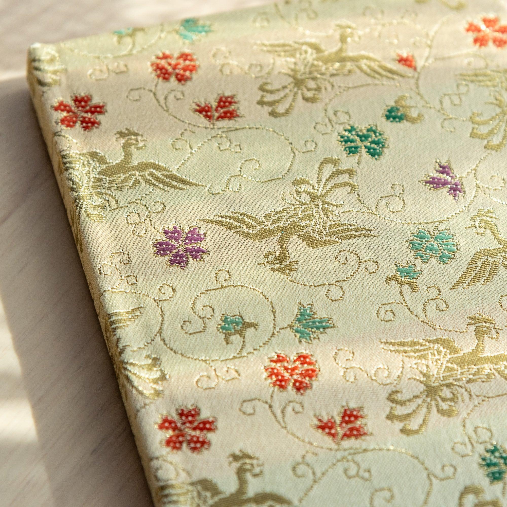 Top edge close-up of Nishijin textile notebook showing cream gold cover with woven phoenix and arabesque scrollwork pattern