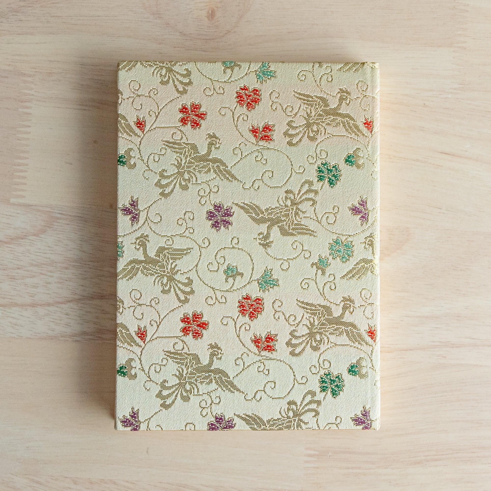 Japanese notebook with pale gold Nishijin woven cover showing phoenix birds and arabesque scrollwork pattern, flat on wood