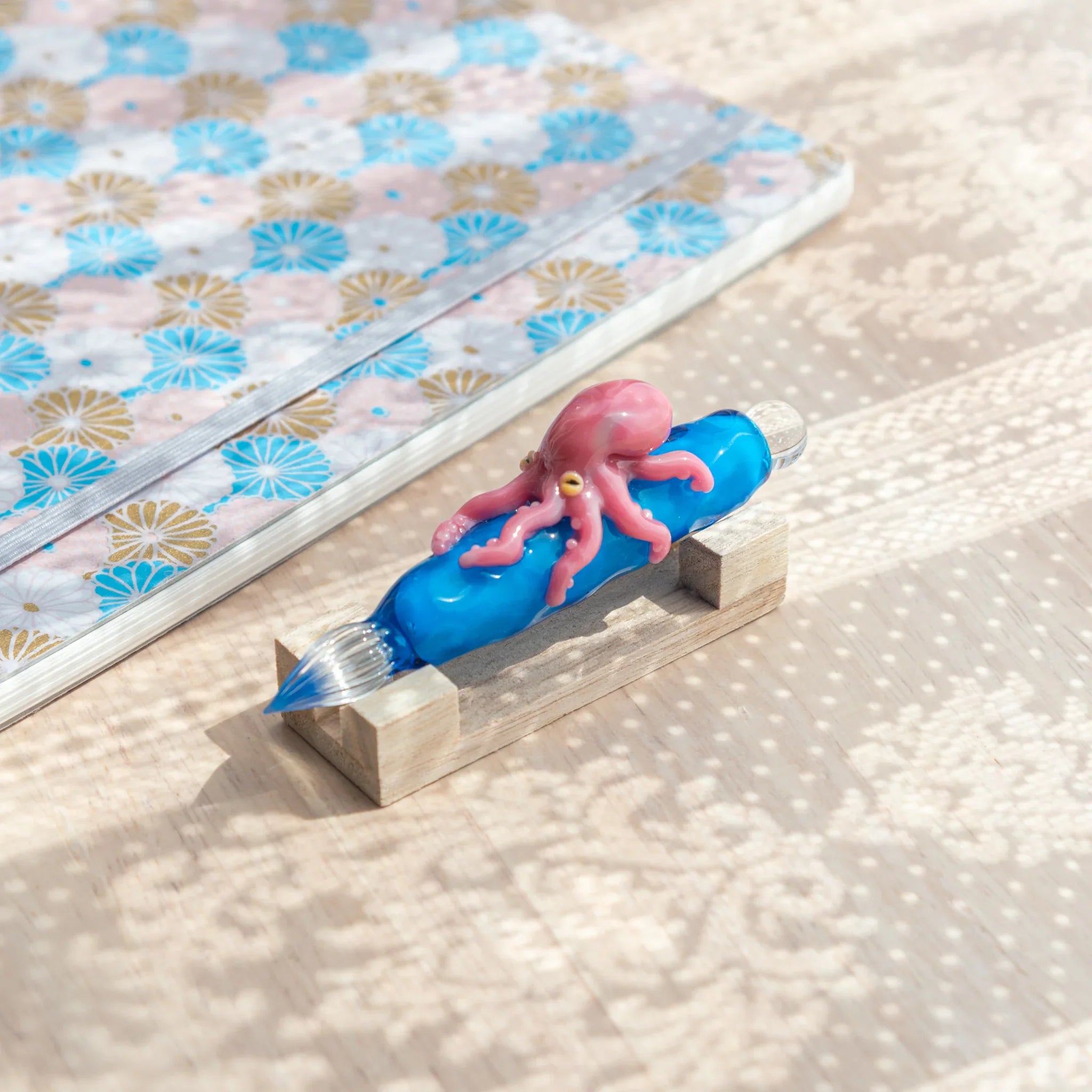 Blue glass Japanese dip pen with pink octopus figurine on natural wood pen rest beside blue floral patterned notebook