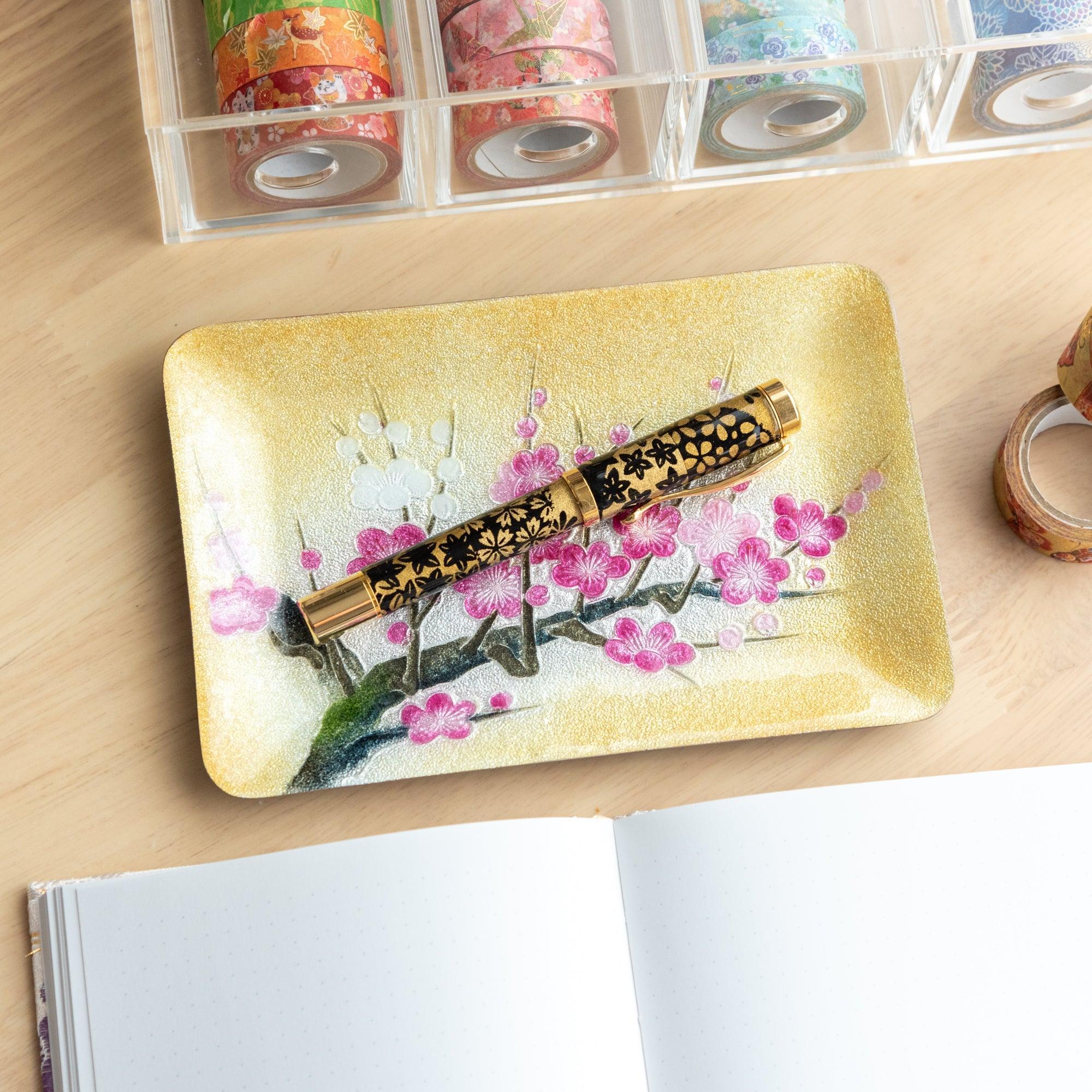 Plum shippo ware gold enamel pen tray on wood desk holding a black floral fountain pen, with washi tape organizer
