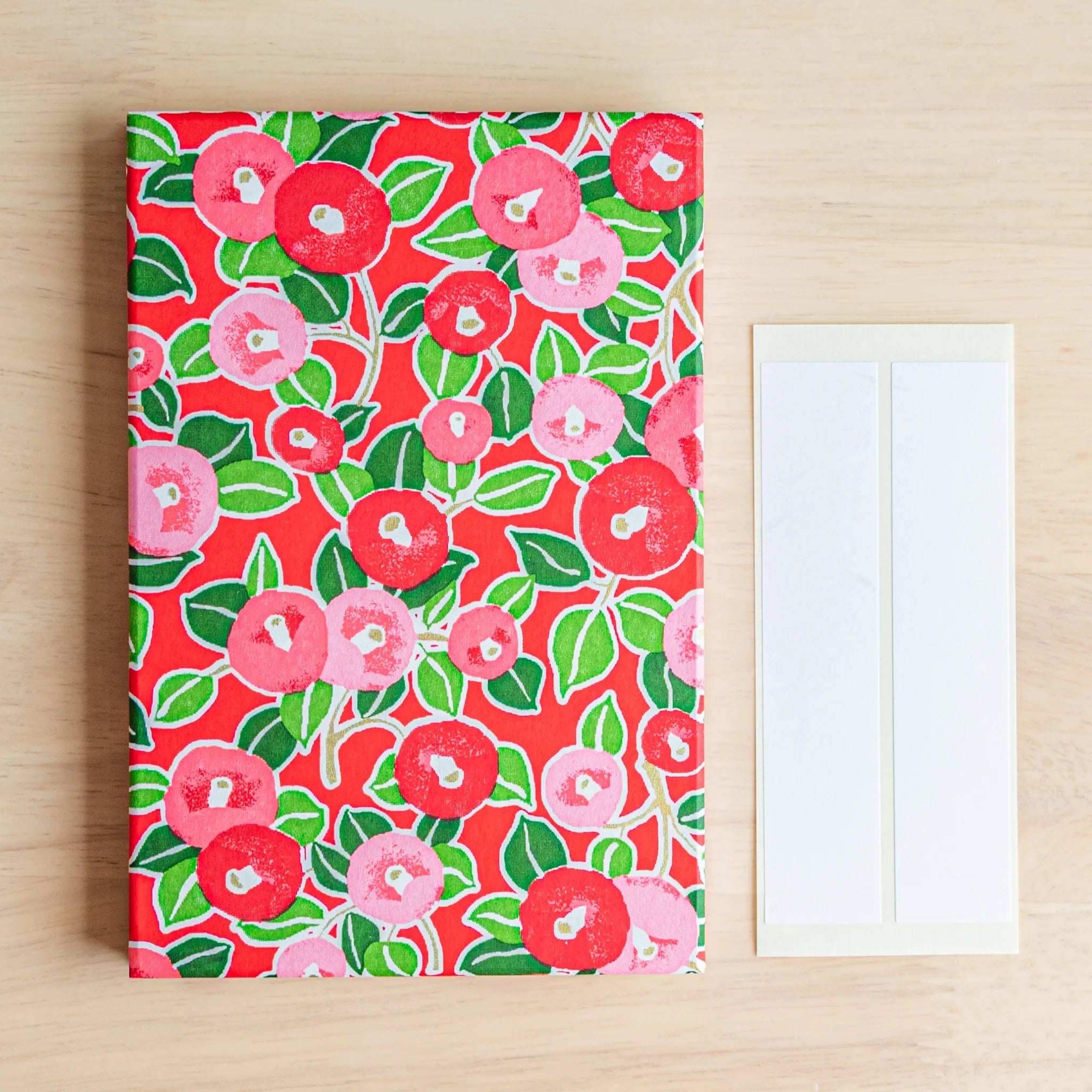 Red Camellia washi goshuincho front cover flat lay on wood surface beside two included cream washi name label strips