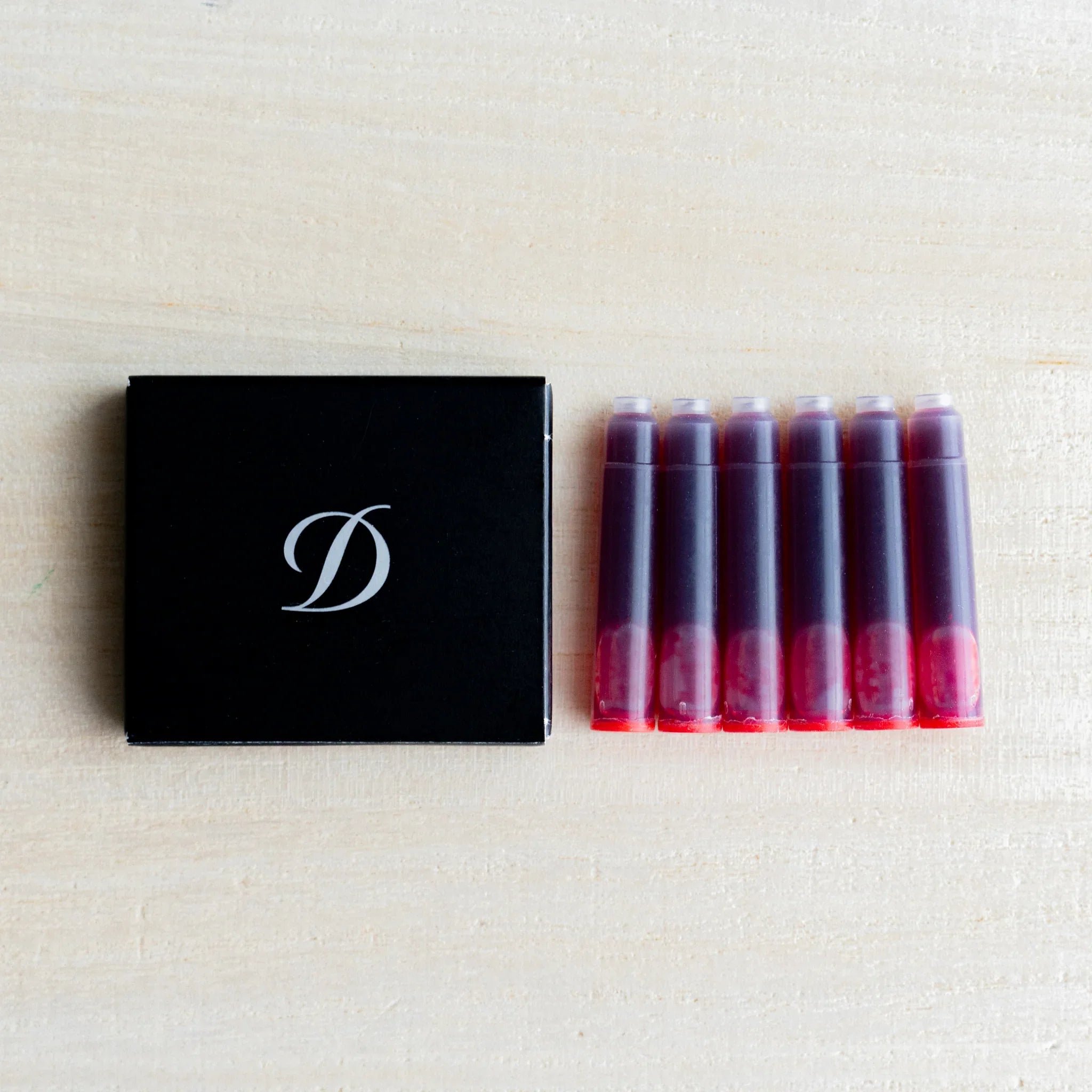 Black S.T. Dupont box with D logo beside six fountain pen ink cartridges holding red ink, on light wood