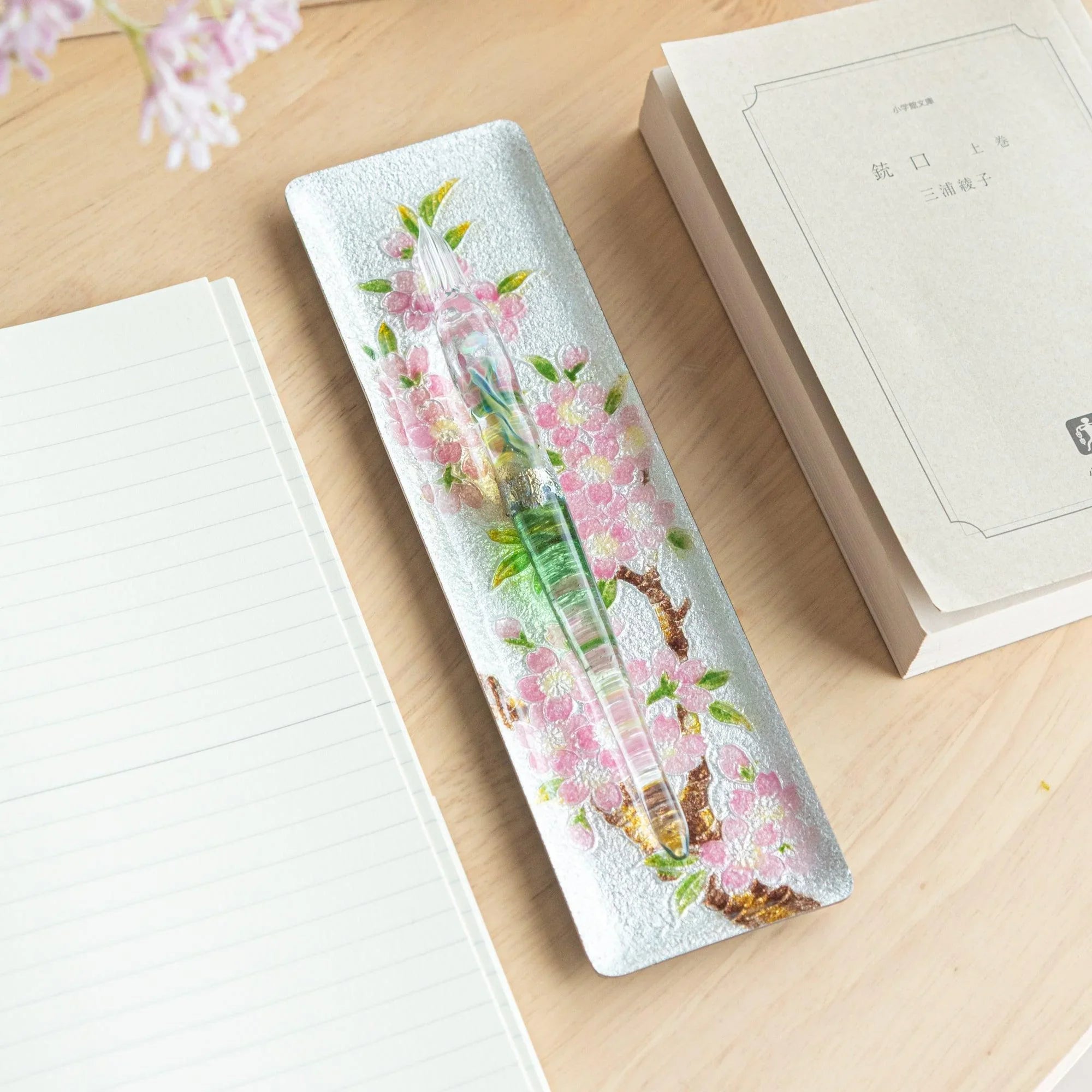 Silver enamel slim pen tray with pink sakura design, glass pen resting on tray, wood desk with notebook and Japanese book