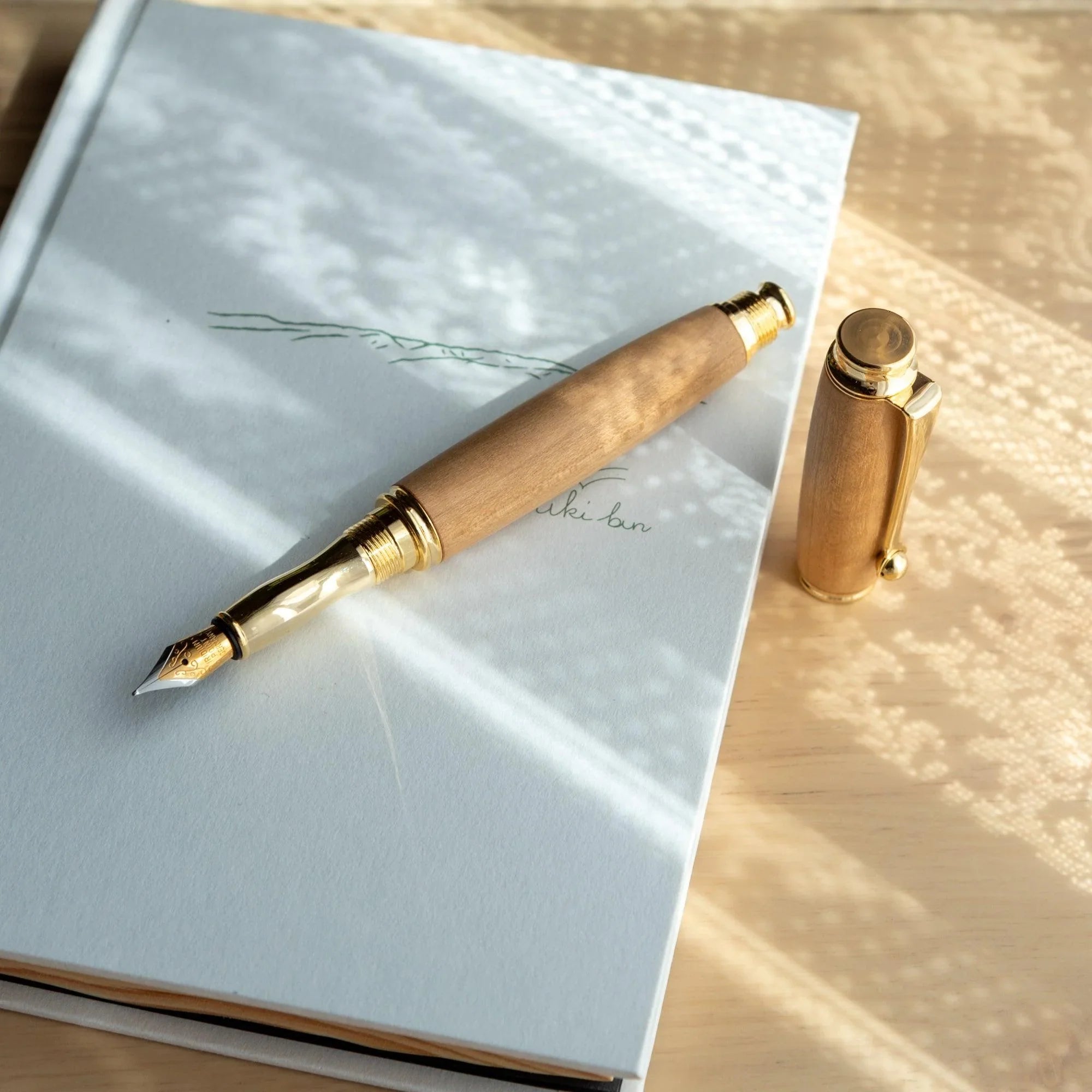 Natural wood Japanese fountain pen open on white notebook with gold nib visible, screw cap set alongside on wood surface