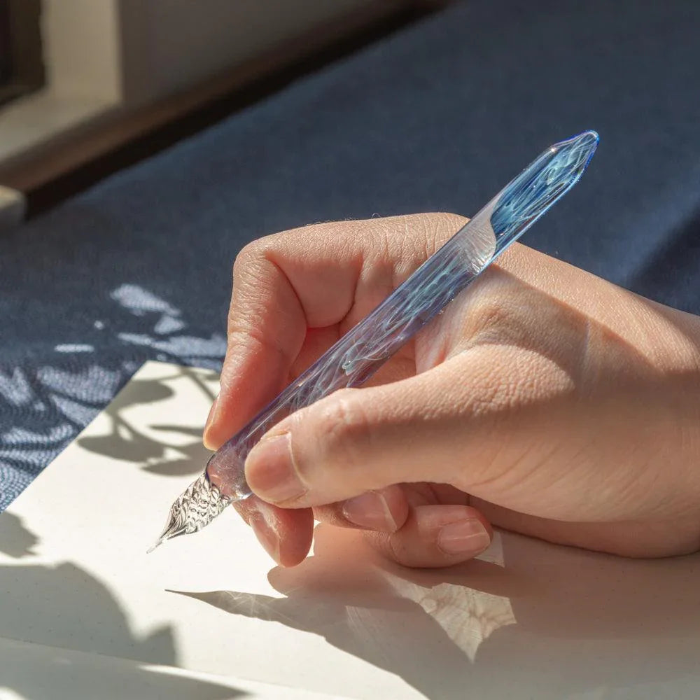Smoke in Azure glass dip pen held in hand with blue-purple glass over white paper with leaf shadow detail
