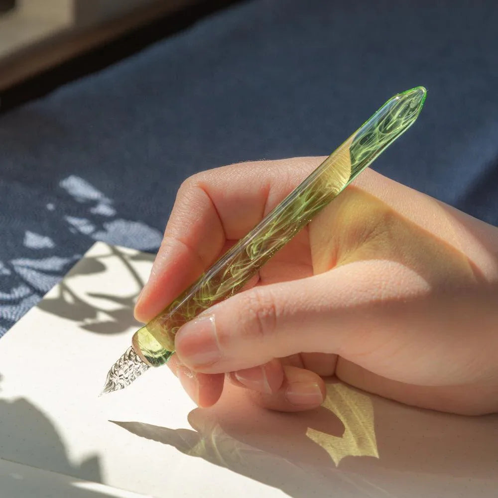 Smoke in Verdure glass dip pen held in hand with green-grey glass over white paper with leaf shadow detail