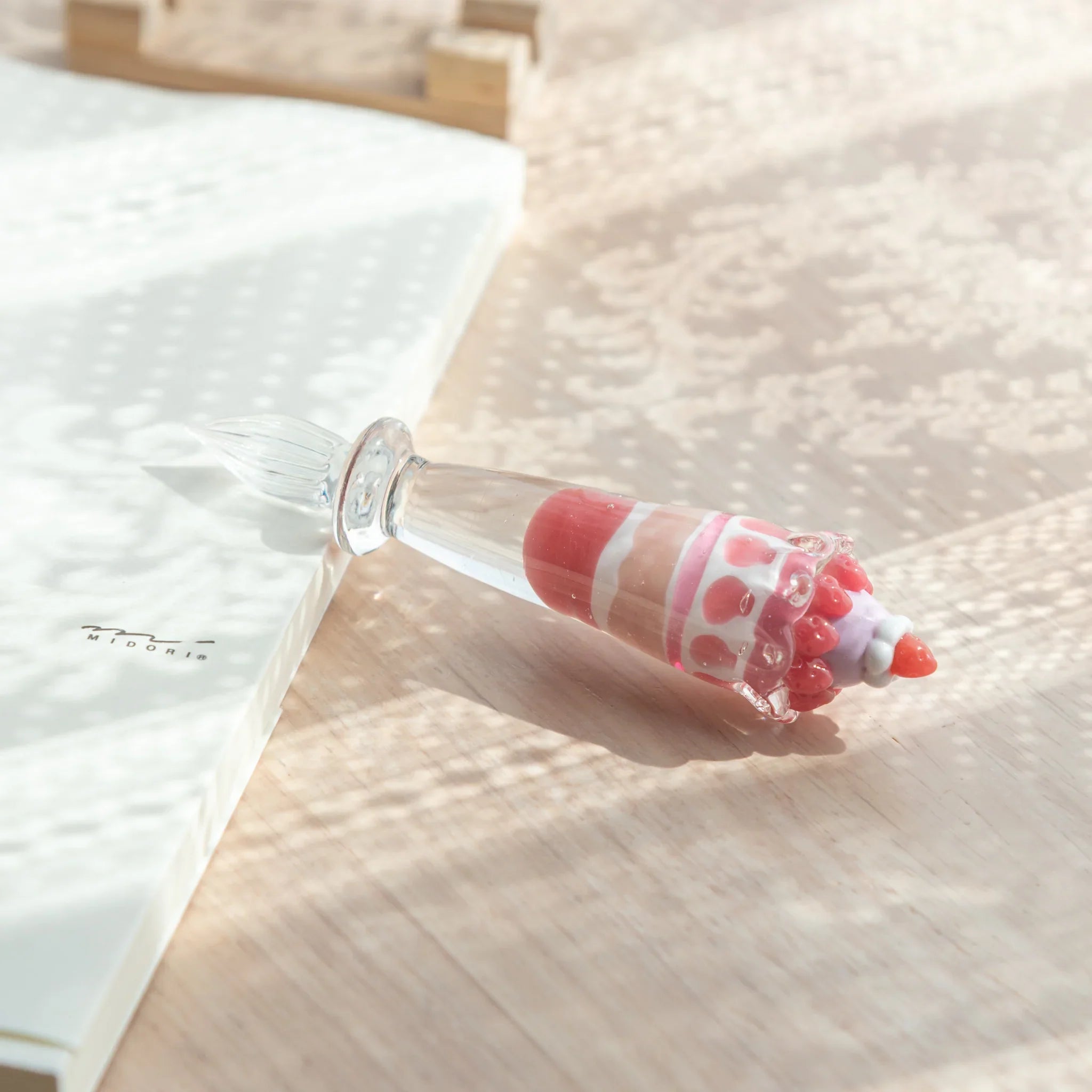 Japanese glass dip pen with strawberry parfait grip in red, pink, and white glass resting on light wood beside a notebook