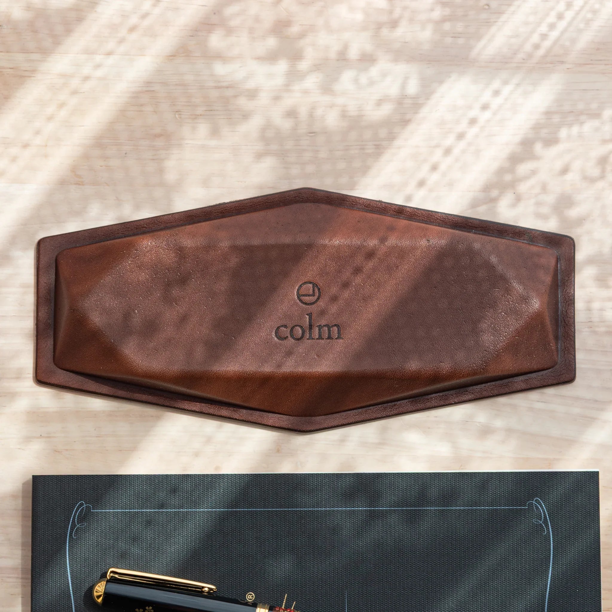Hexagonal slim pen tray upside down showing brown leather base with embossed colm logo and hexagonal outline on wood surface