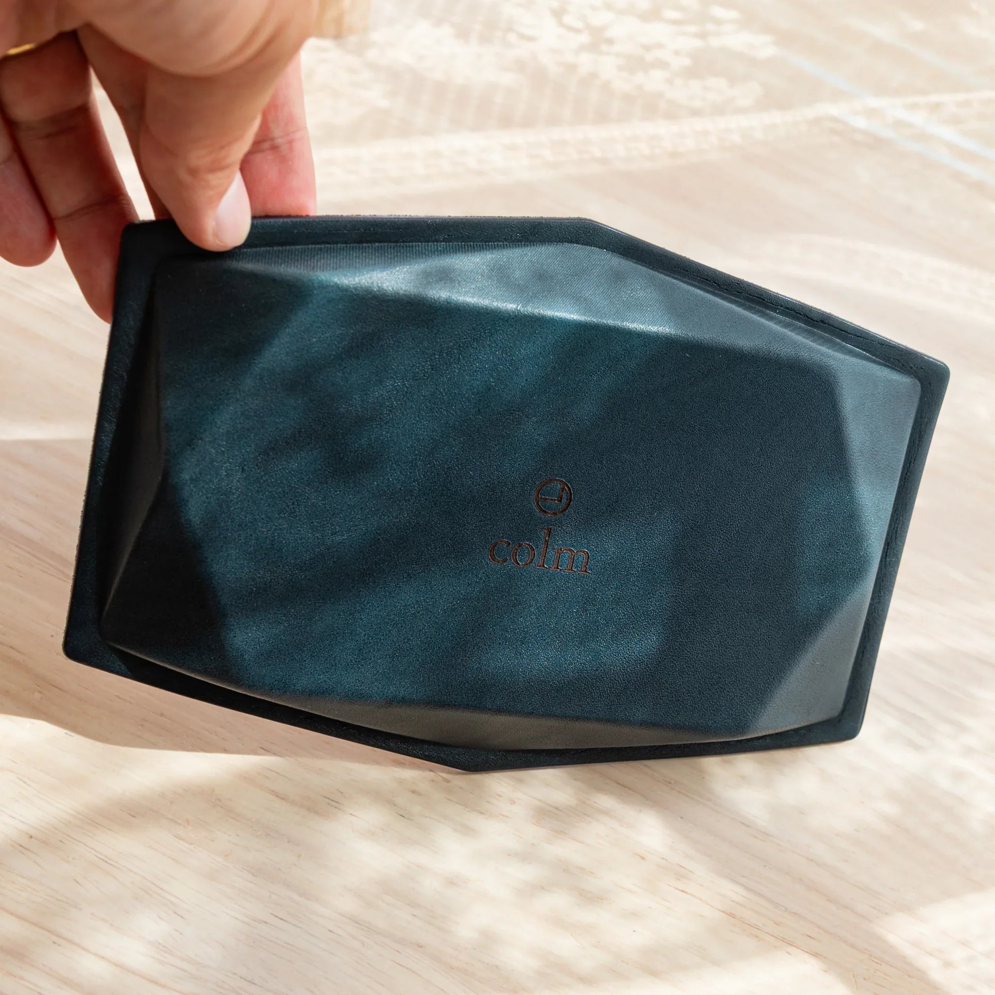 Hand tilting blue-gray suede wide hexagonal pen tray to reveal dark teal suede base with embossed colm logo