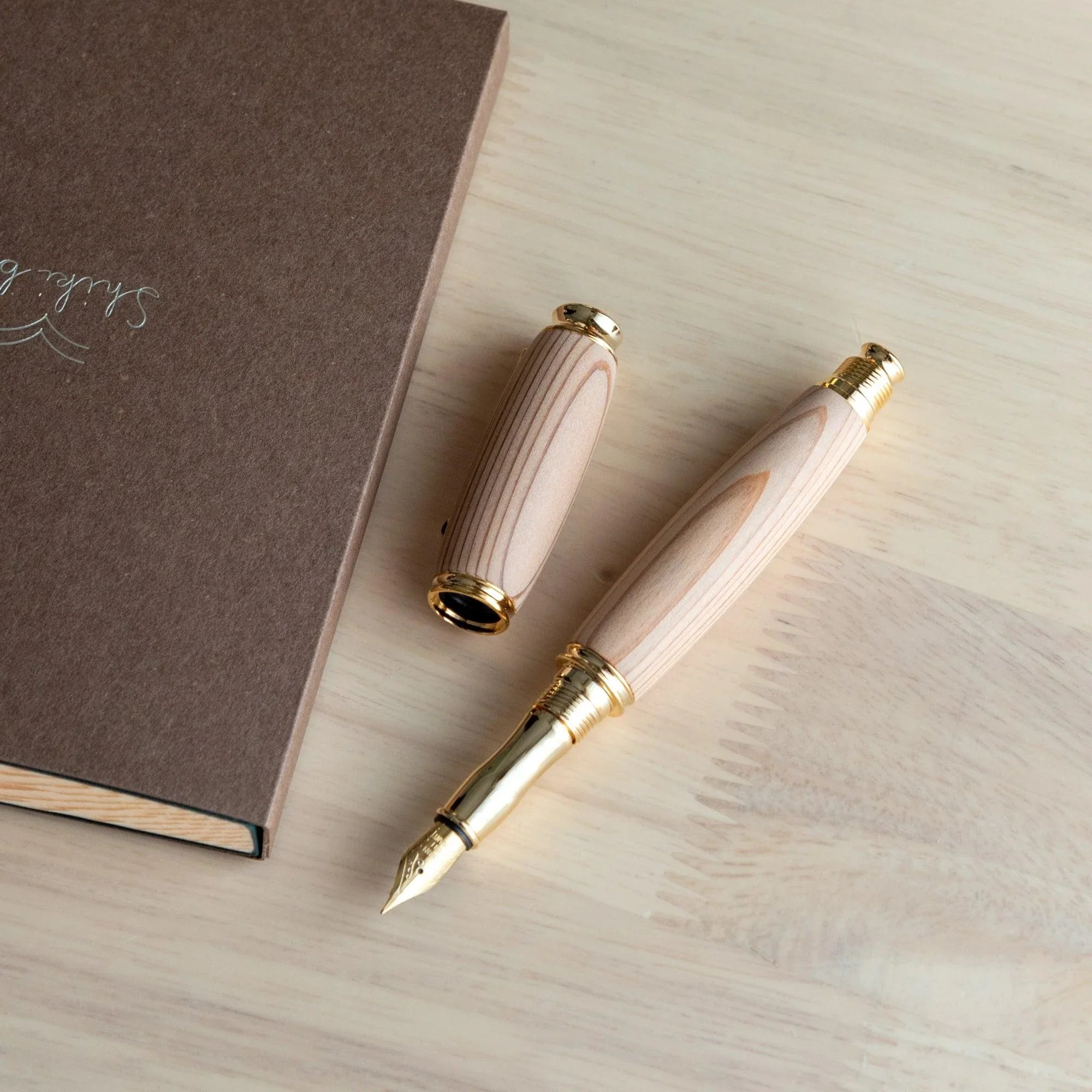 Cedar wood Japanese fountain pen uncapped, cap beside body on light wood desk, gold nib visible, brown notebook at left