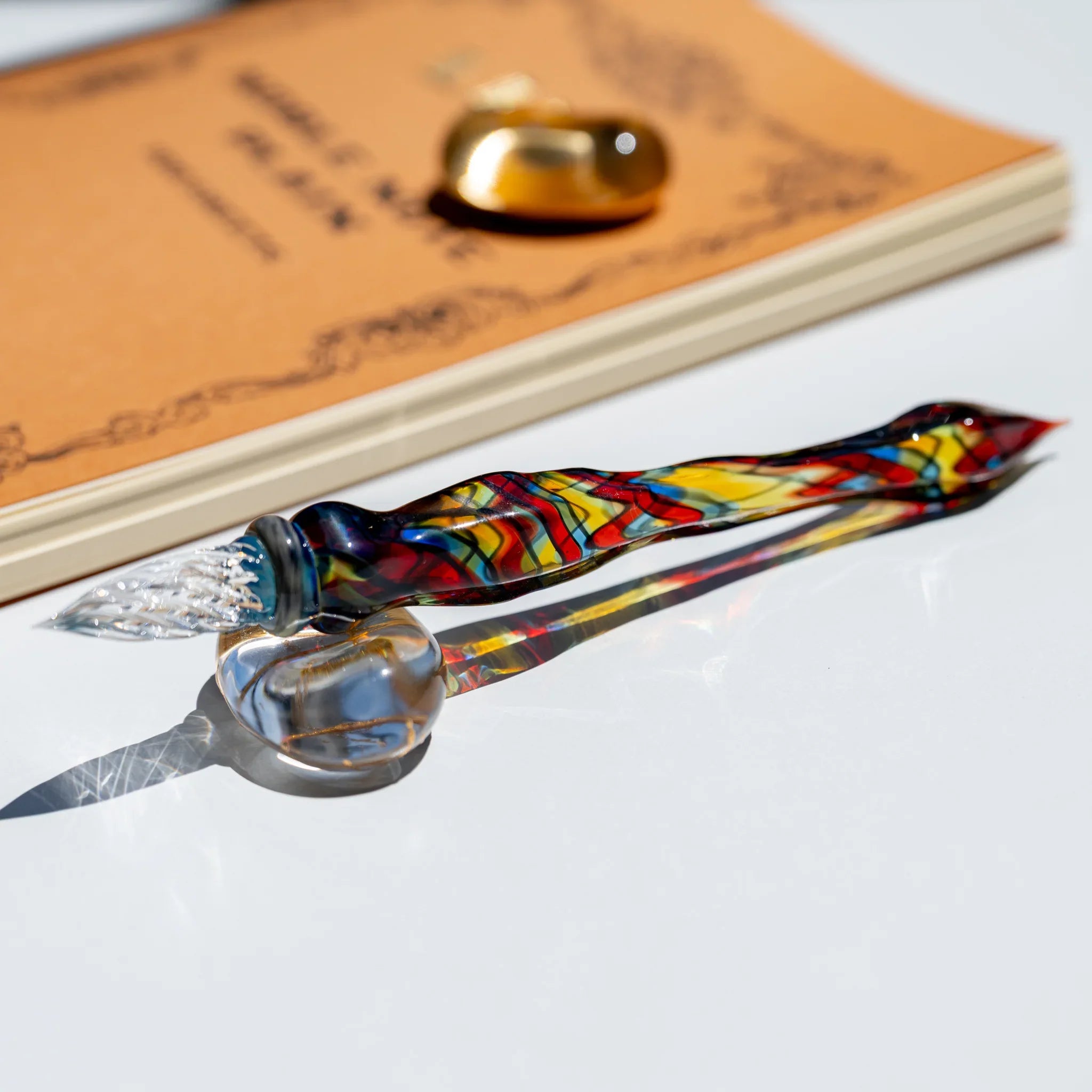 Full glass dip pen with clear nib resting on a round glass rest beside a kraft notebook, mosaic body in red, yellow and blue
