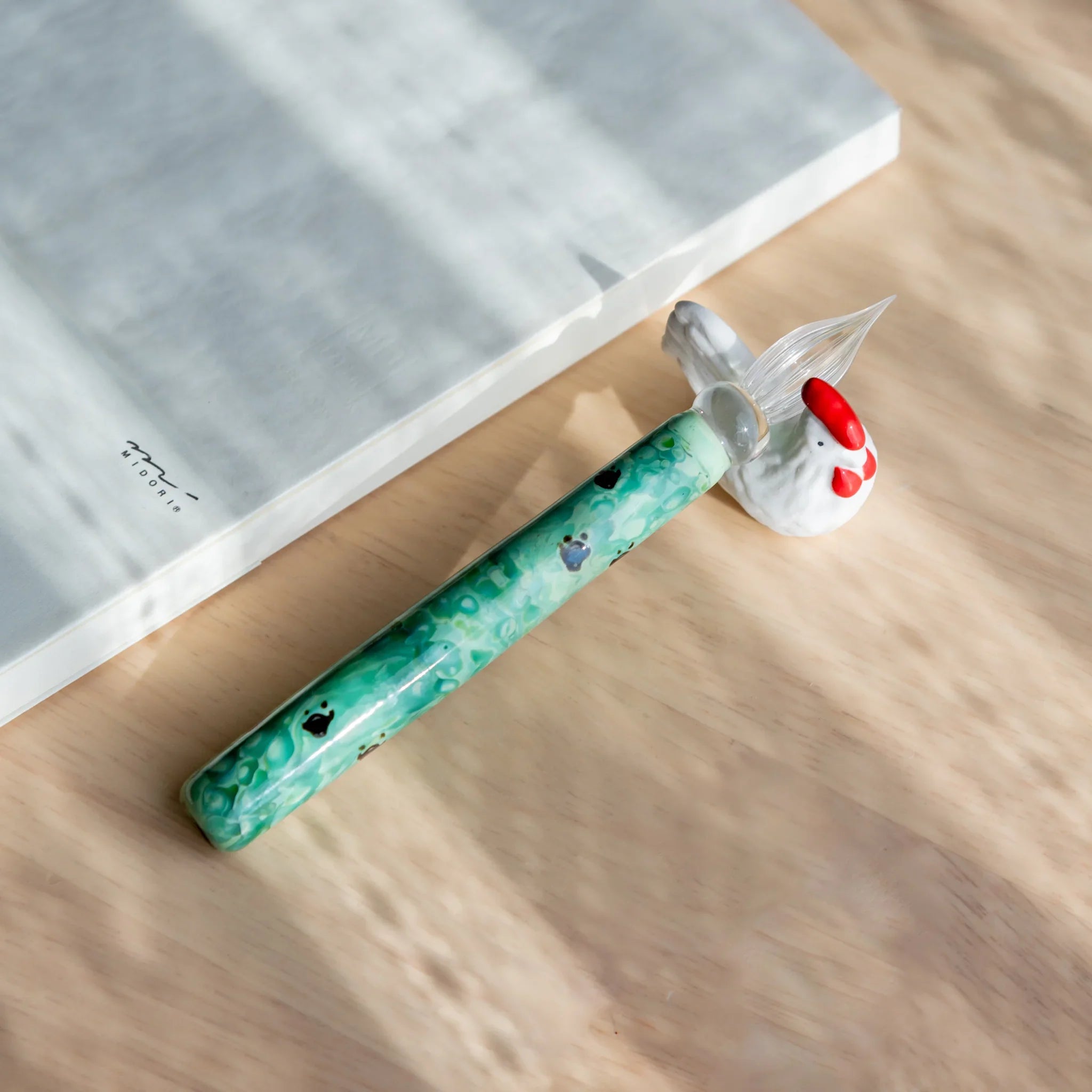 White ceramic chabo rooster pen rest holding a turquoise mottled glass pen on wood surface beside an open Midori notebook