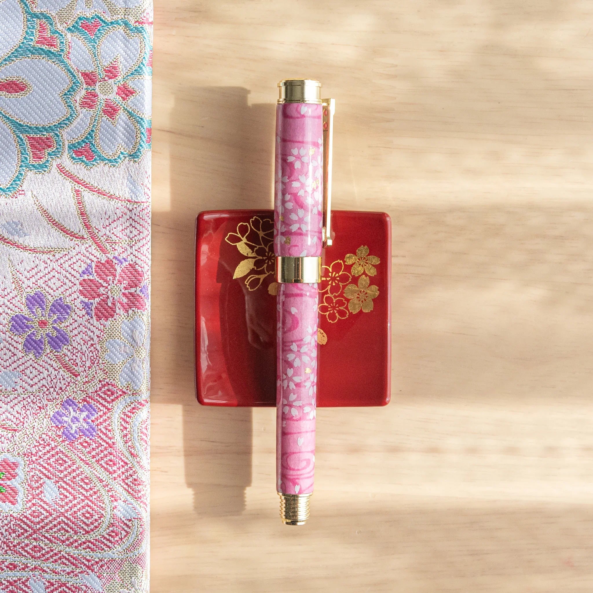 Pink Mino washi Japanese fountain pen with white sakura print and gold trim, resting on a red lacquer pen tray