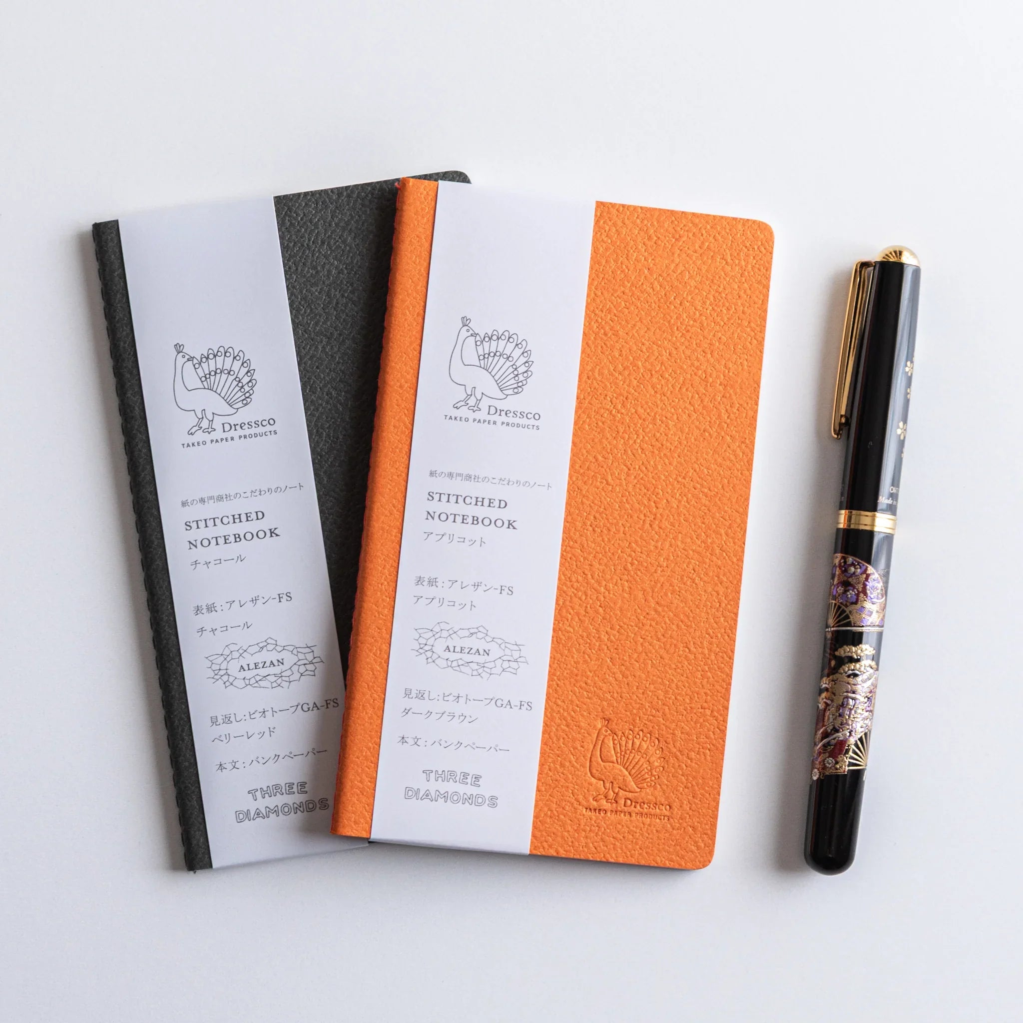 Three A6 Dressco stitched notebooks with charcoal gray and orange fabric covers displayed with decorative fountain pen