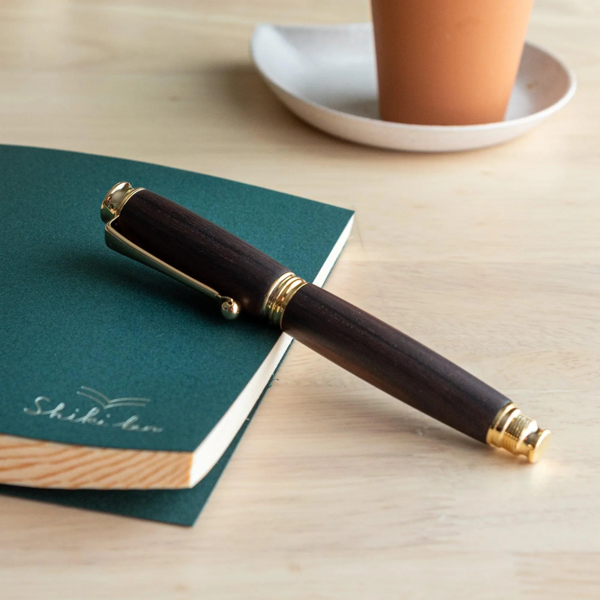 Dark ebony wood Japanese fountain pen capped on a green Shiki An notebook on a wood desk, terracotta cup in background