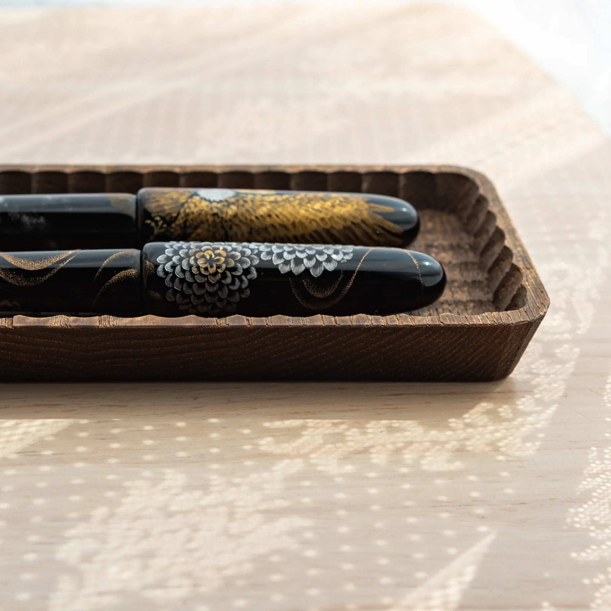 Charcoal Japanese pen tray at a low side angle with two black fountain pens in gold feather and chrysanthemum designs
