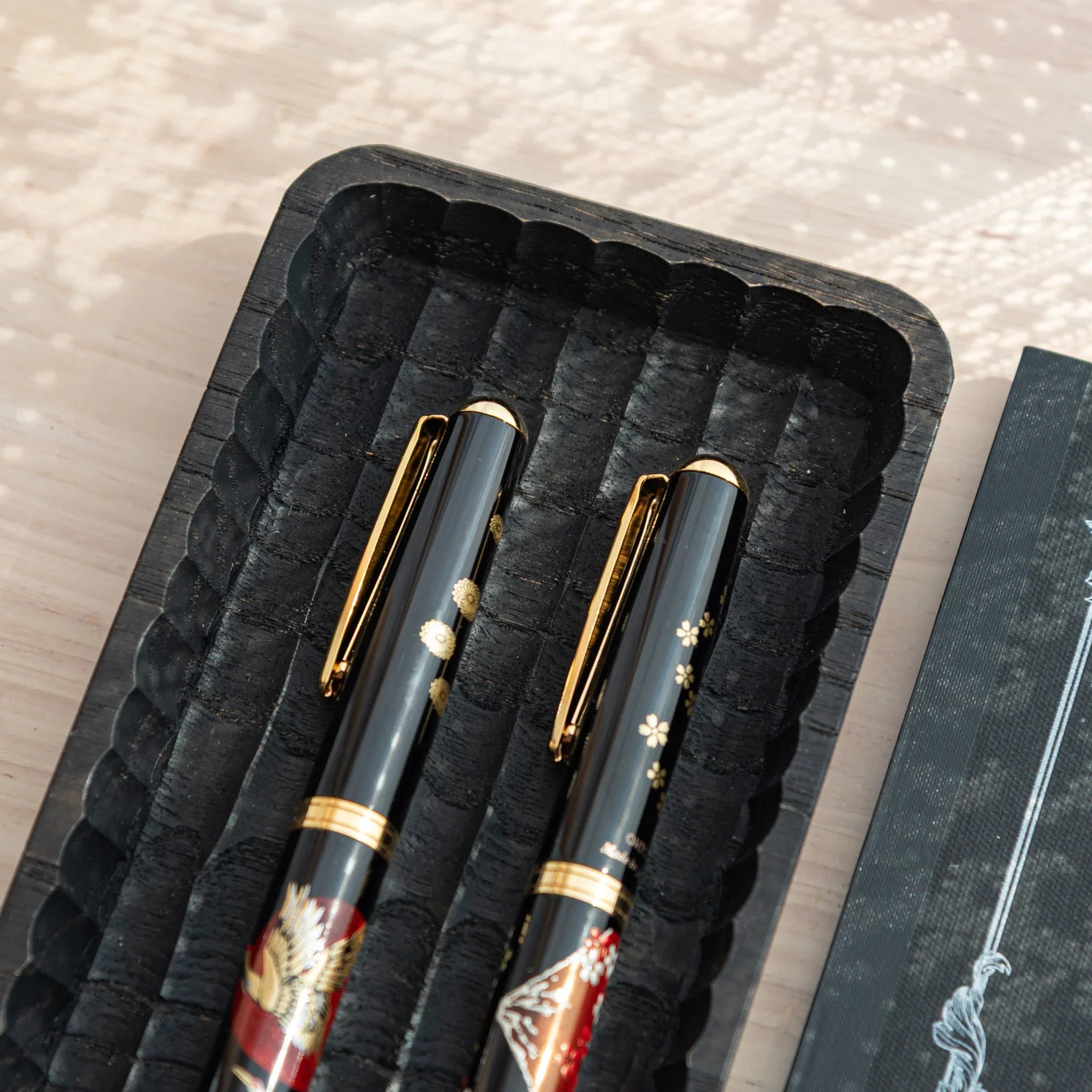 Black Japanese pen tray close-up at angle with two fountain pens in crane and Mt. Fuji sakura designs, notebook beside