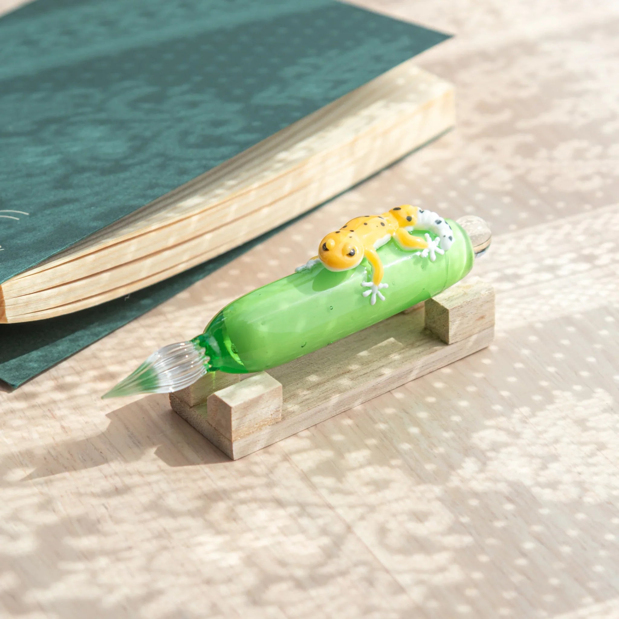 Green glass dip pen with yellow spotted leopard gecko figure on barrel on natural wood pen stand, teal notebook in background