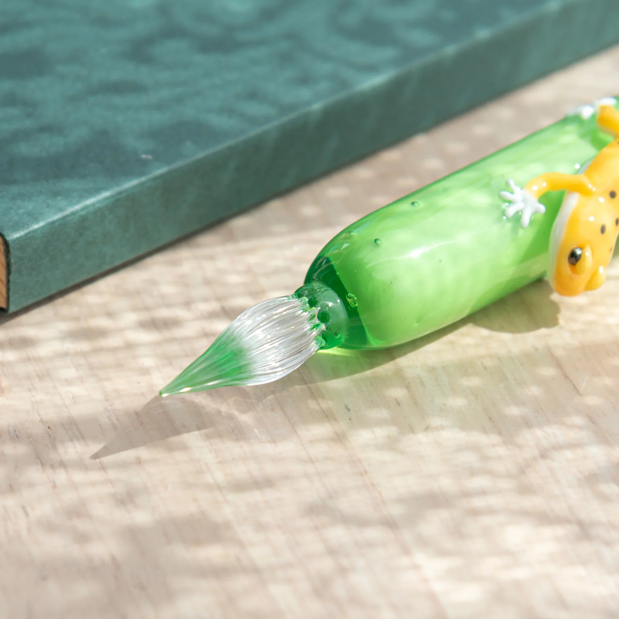 Green glass dip pen on wood surface showing ribbed clear glass nib and yellow spotted leopard gecko, teal notebook behind
