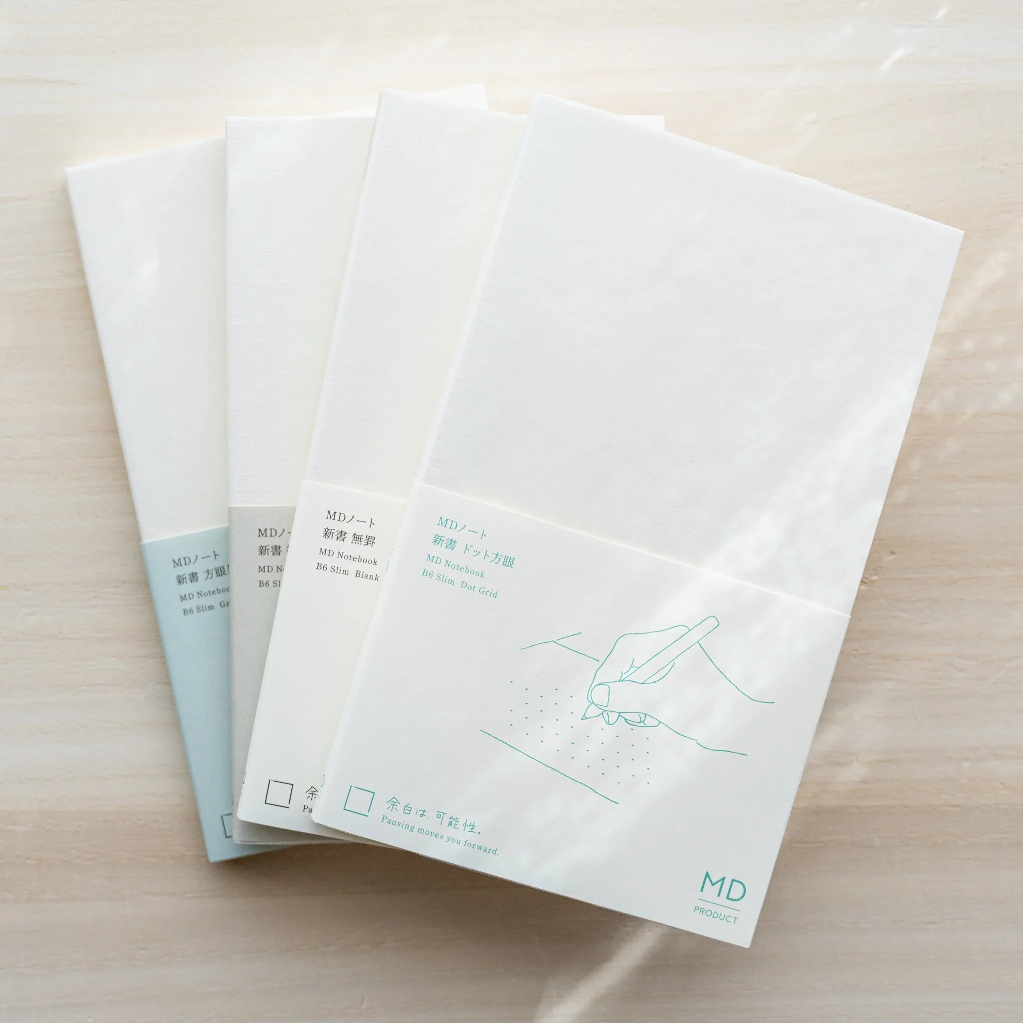 Three Midori MD Notebook B6 Slim notebooks arranged fan-style on wood surface, white covers with teal hand illustration