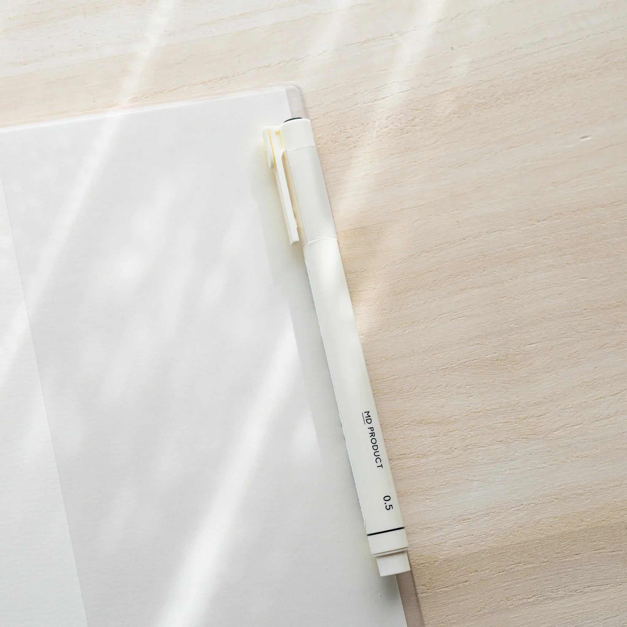 White MD PRODUCT 0.5mm pen resting diagonally across a clear PVC Midori MD Notebook Cover