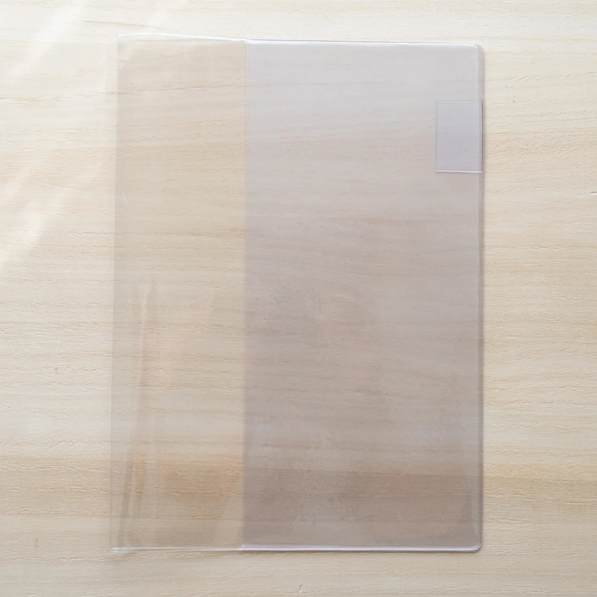 Clear PVC Midori MD Notebook Cover alone on a wood surface, transparent with a white bookmark tab
