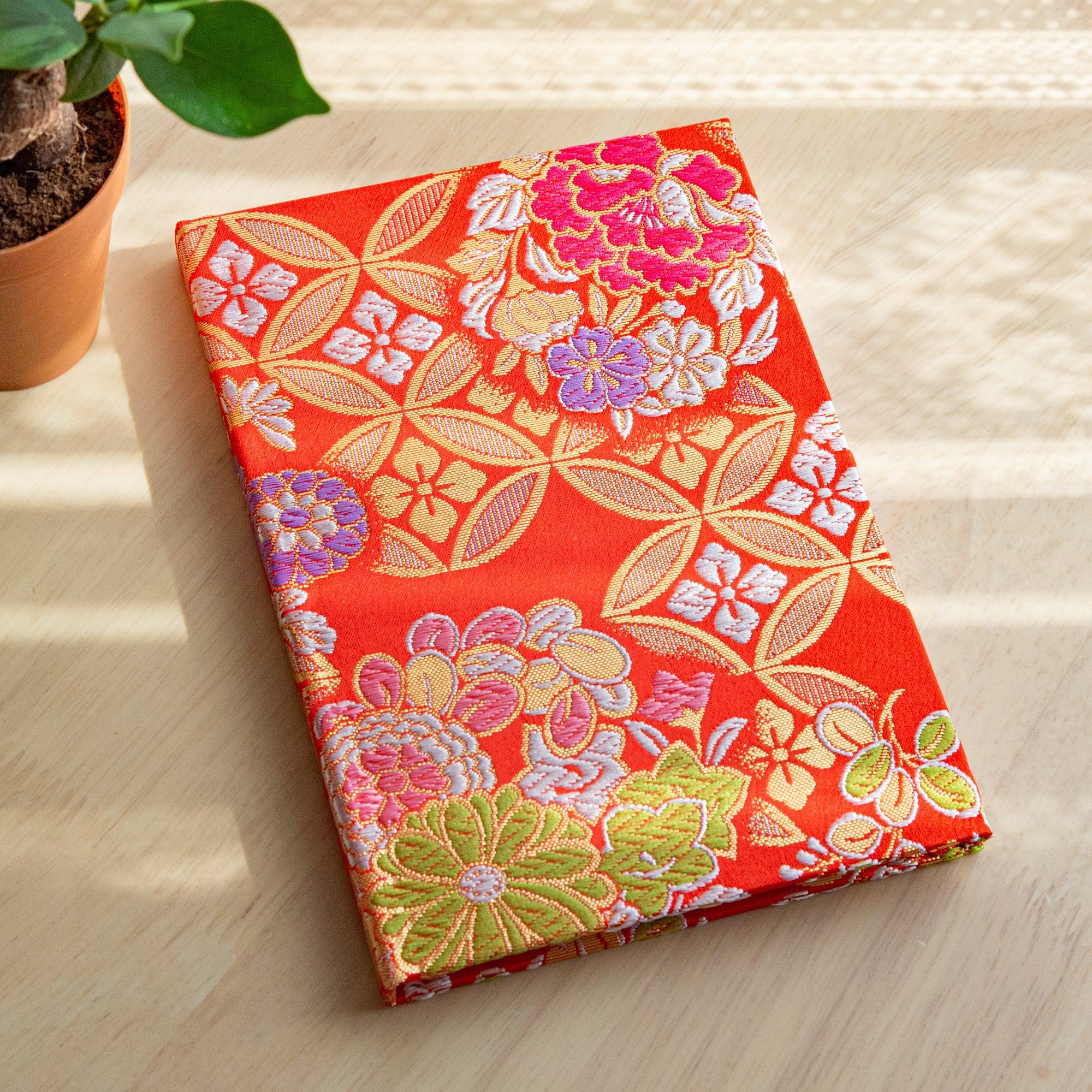 Japanese notebook with orange-red Nishijin woven textile cover in shippo floral pattern, angled on wood with a plant