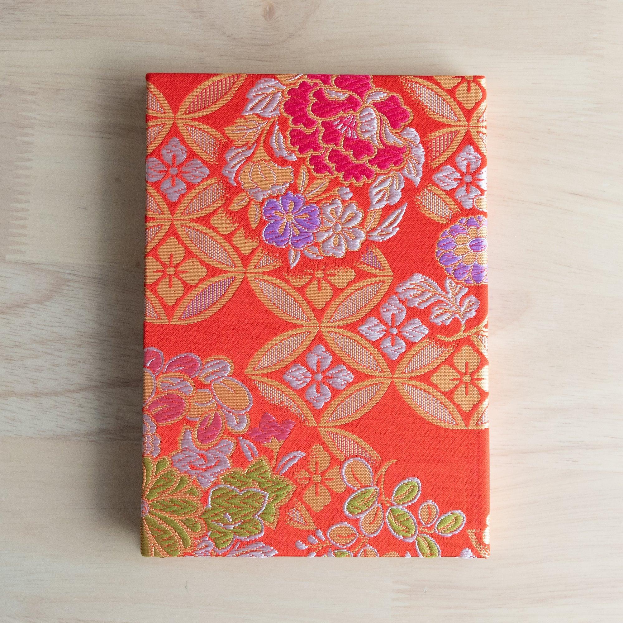 Nishijin textile Japanese notebook with orange-red shippo cover showing magenta peonies and purple flowers, flat on wood