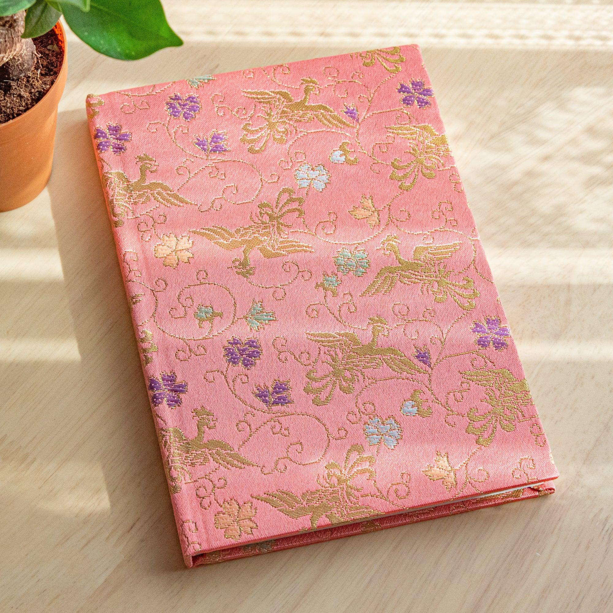 Japanese notebook with pink Nishijin woven cover showing gold phoenix and arabesque, angled on white surface with plant