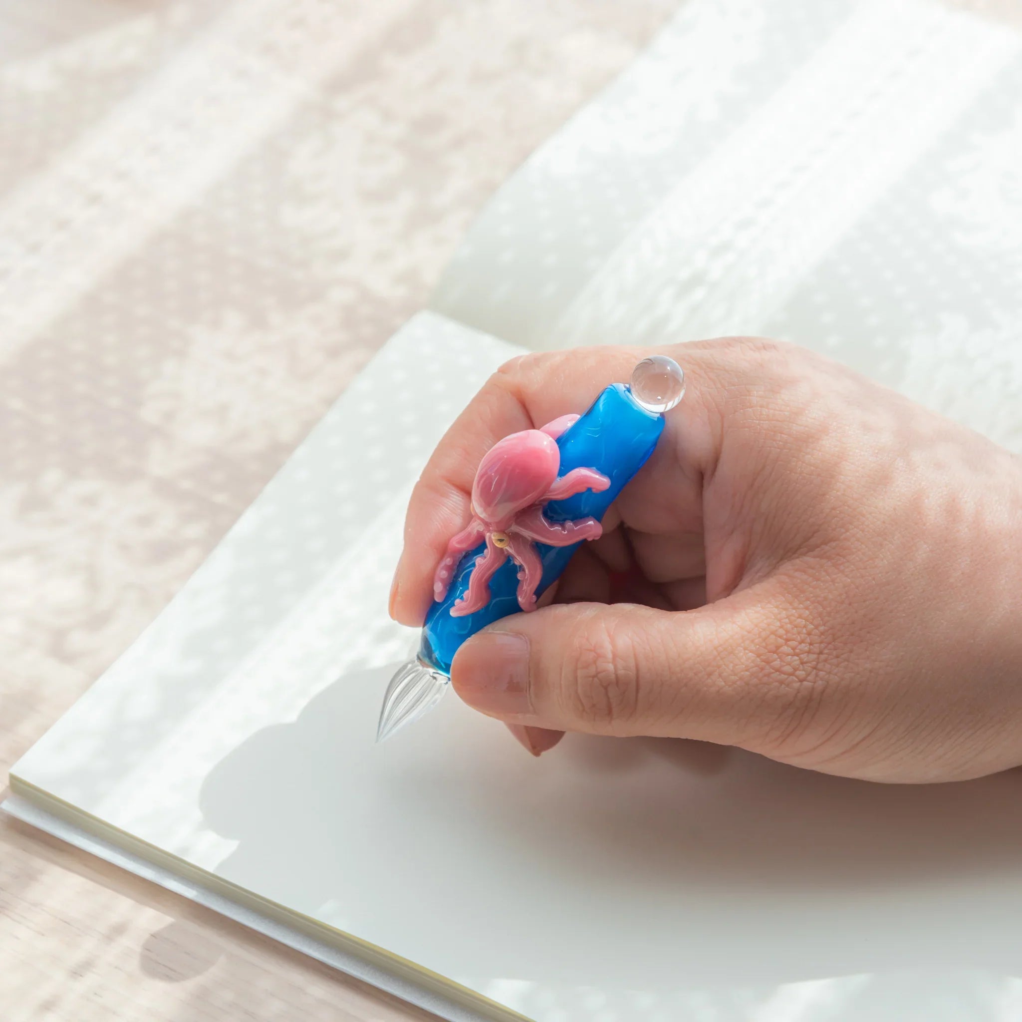 Hand holding blue glass Japanese dip pen with pink octopus figurine, clear spiral nib over open white notebook