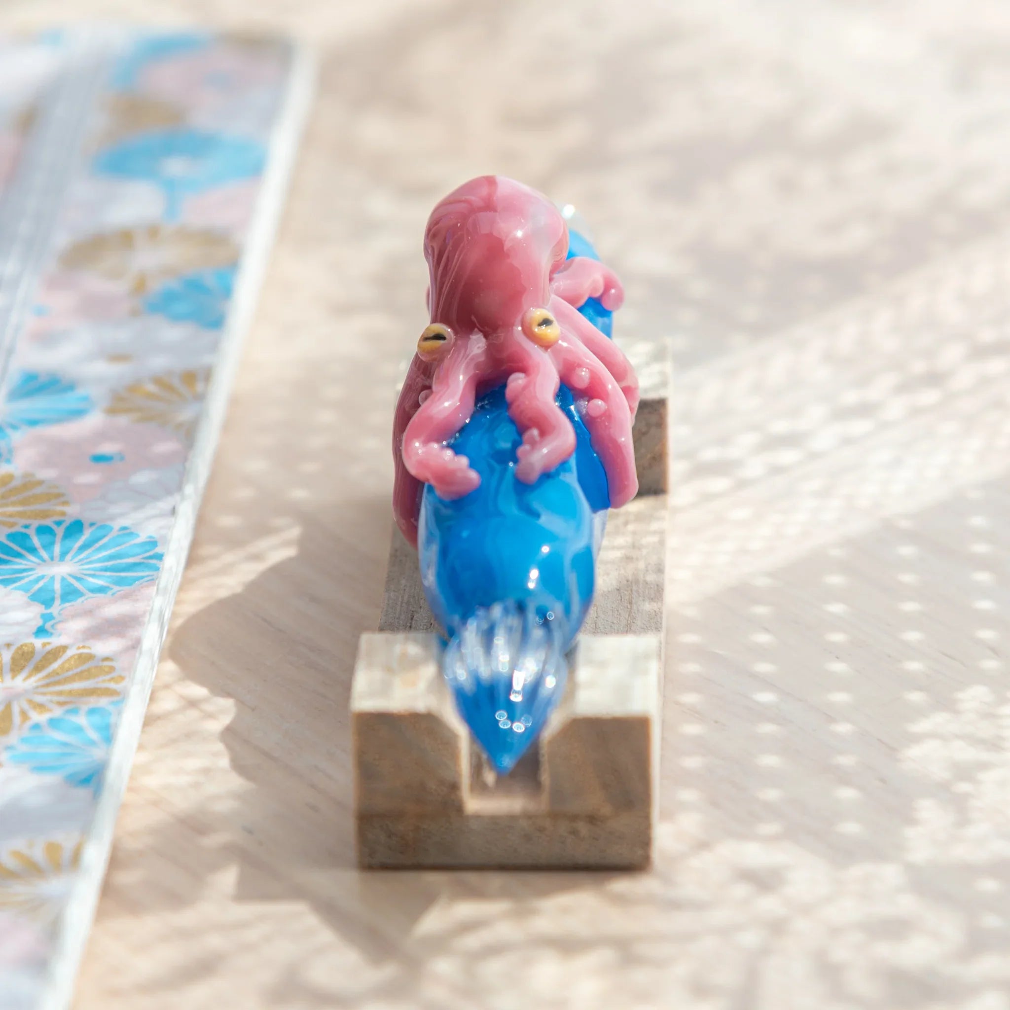 Blue glass Japanese dip pen with pink octopus figurine on natural wood U-shaped pen rest, viewed from above
