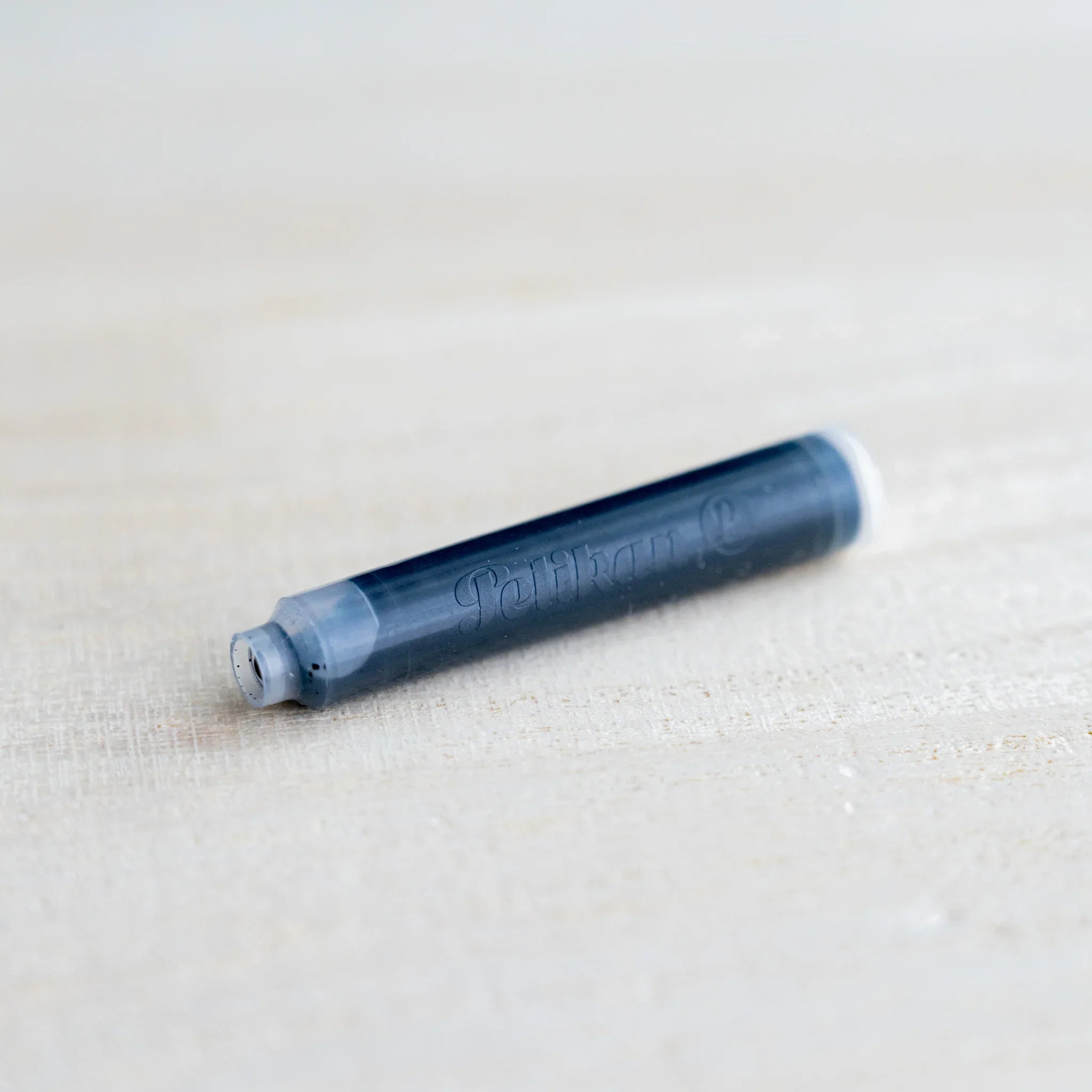 Single Pelikan fountain pen ink cartridge in dark ink, embossed Pelikan, close view on light wood