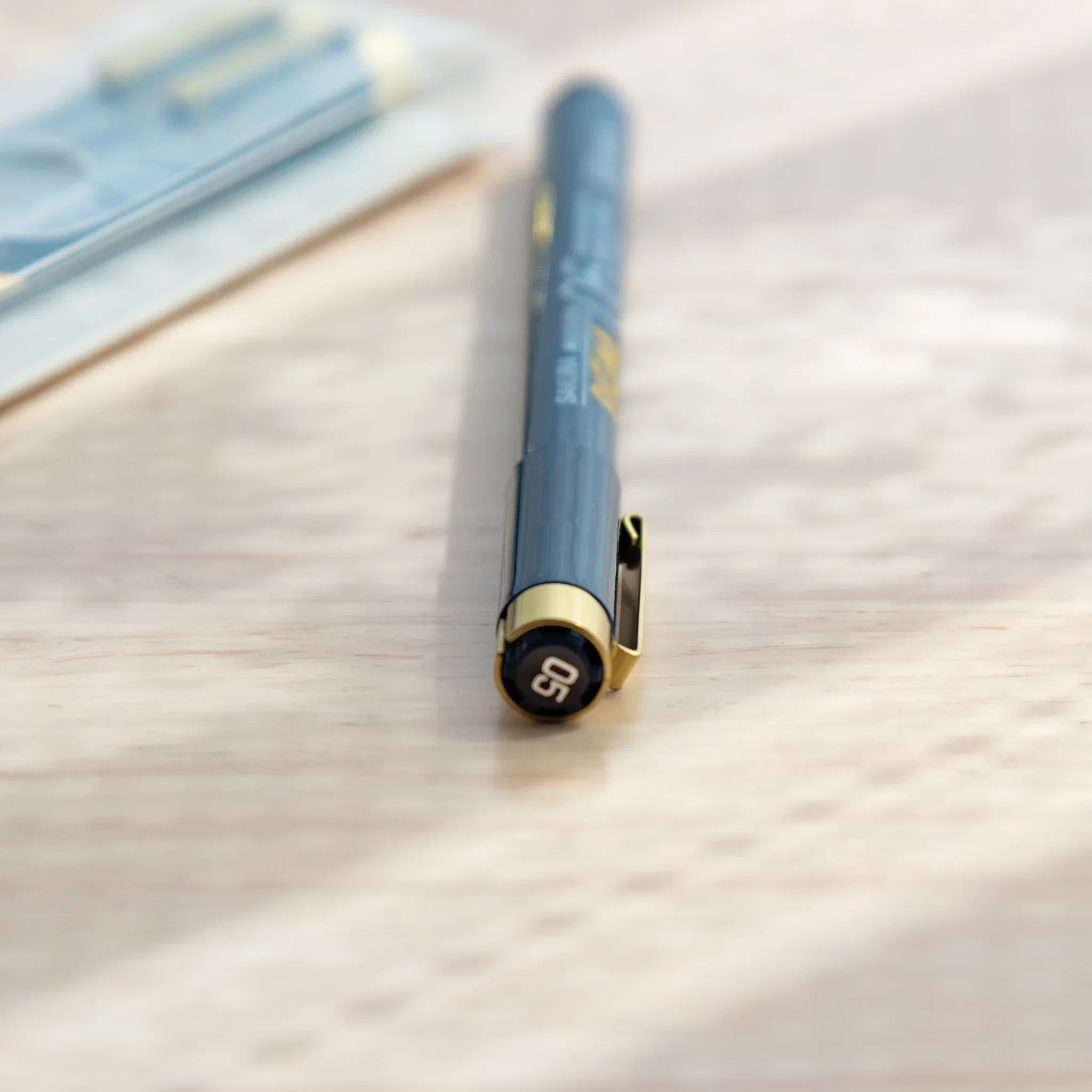 Sakura Pigma Micron 05 fineliner cap end close-up showing size number in gold circle, blue-gray barrel, and blurred packaging