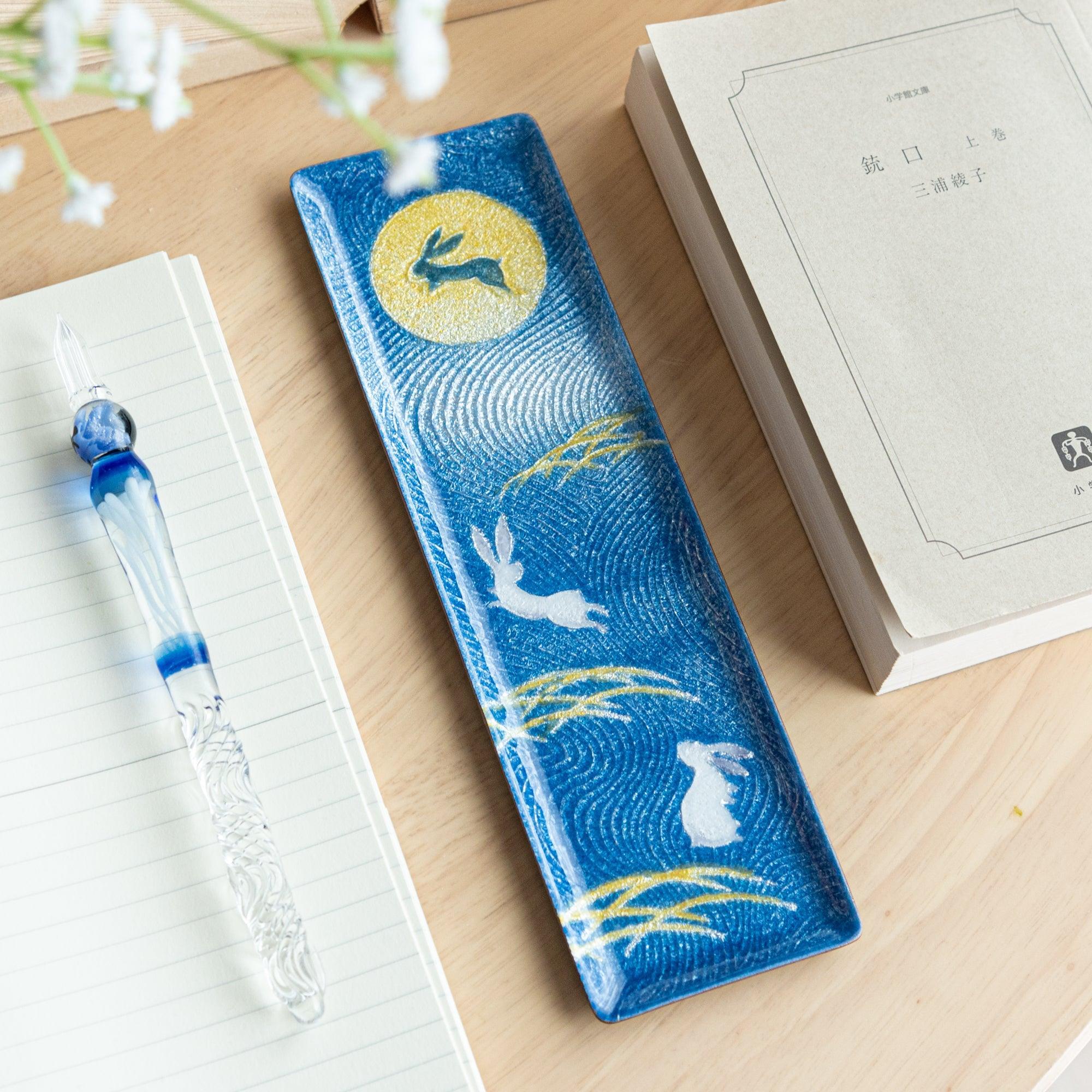 Dark blue enamel slim pen tray with white rabbits and gold moon design, on wood desk with blue glass pen on lined notebook