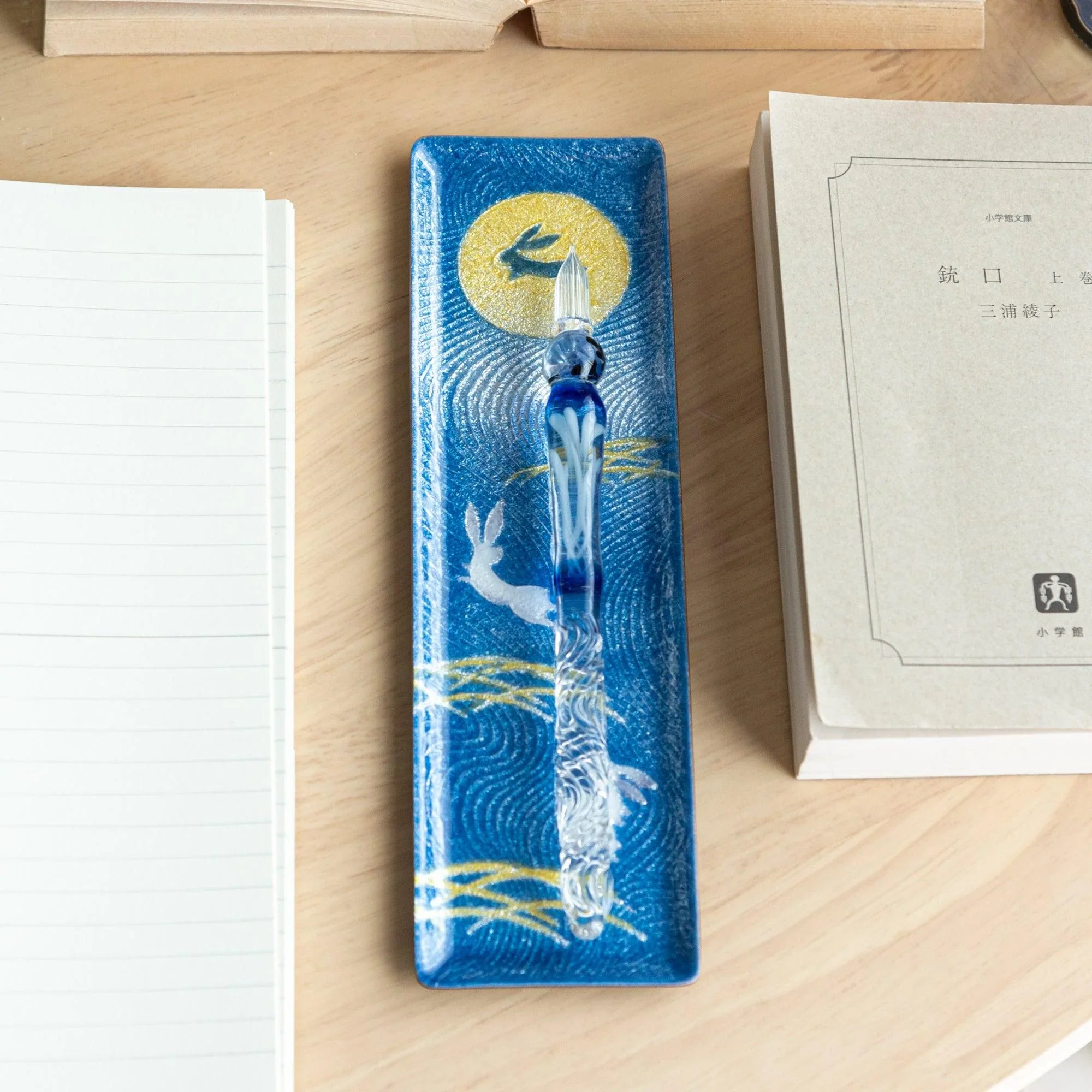 Dark blue enamel slim pen tray with rabbit and gold moon design, overhead view, blue glass pen on tray, notebook to left