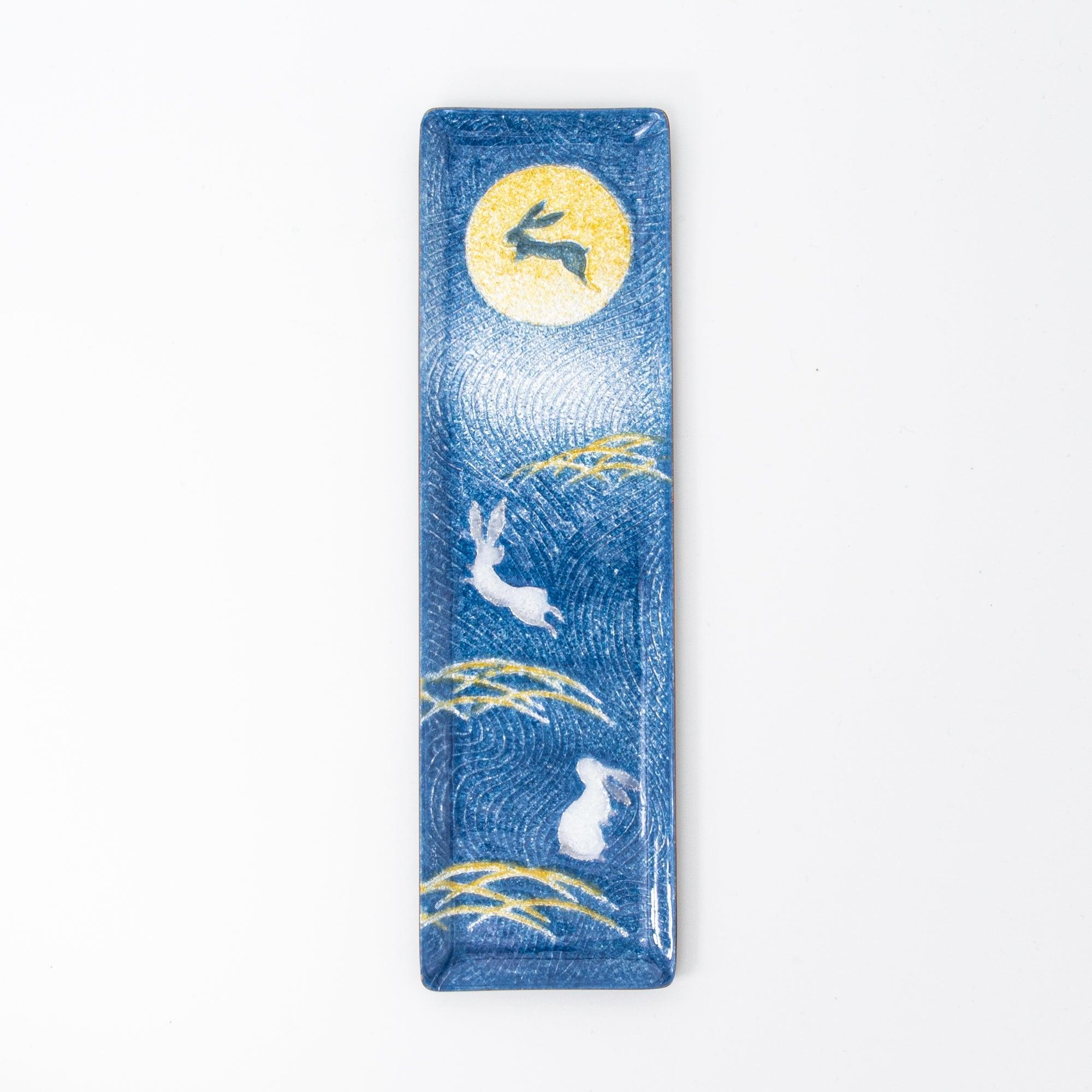 Dark blue enamel slim pen tray showing gold moon, leaping white rabbit, and gold pampas grass, overhead on white background
