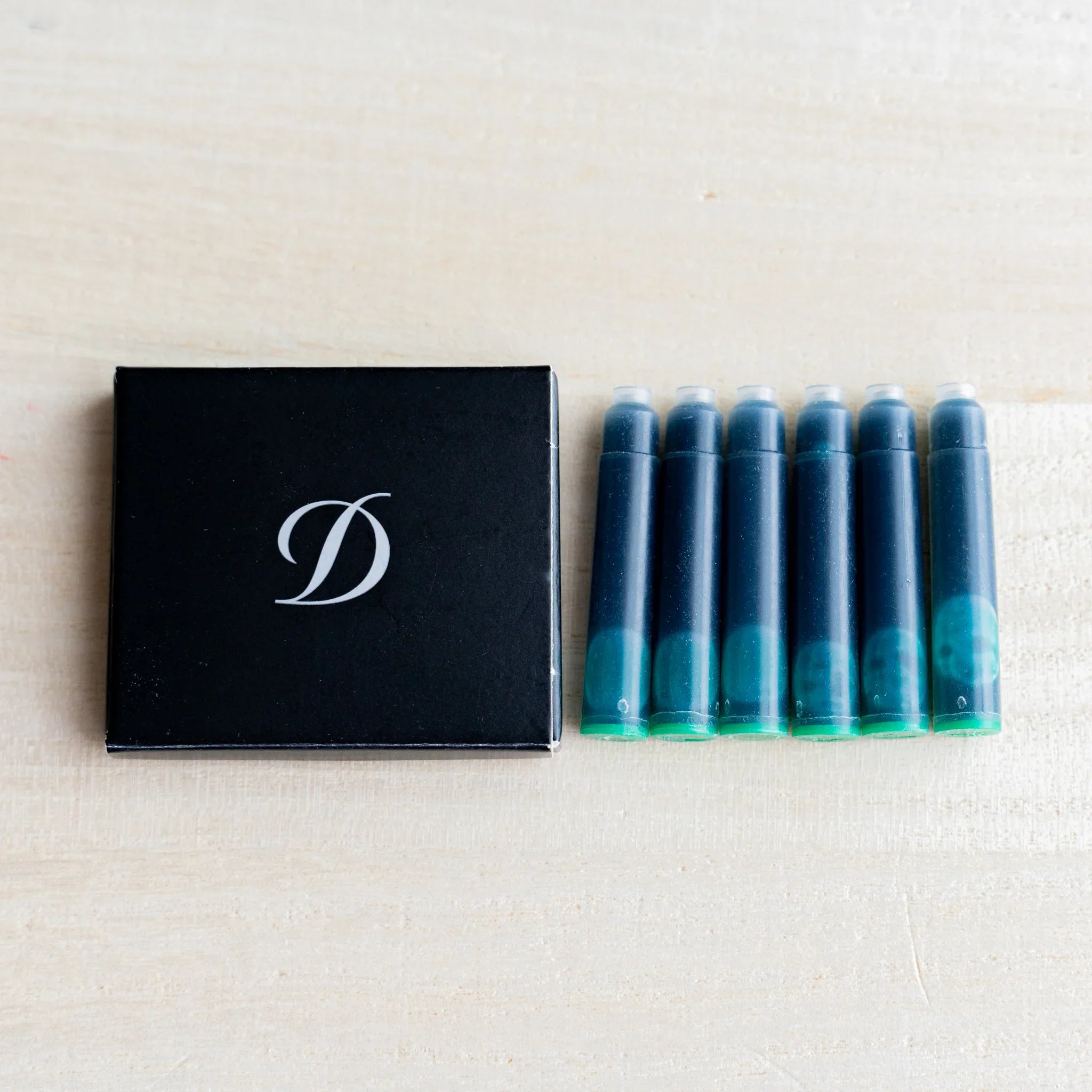 Black S.T. Dupont box with D logo beside six fountain pen ink cartridges holding green ink, on light wood