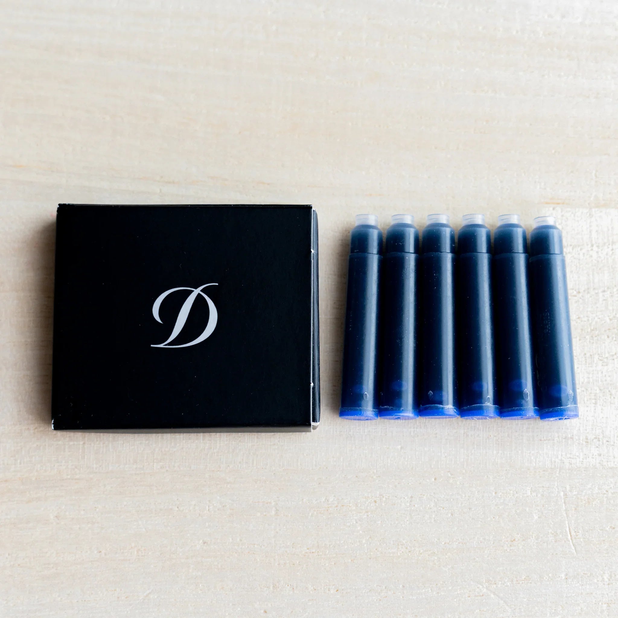 Black S.T. Dupont box with D logo beside six fountain pen ink cartridges holding blue ink, on light wood