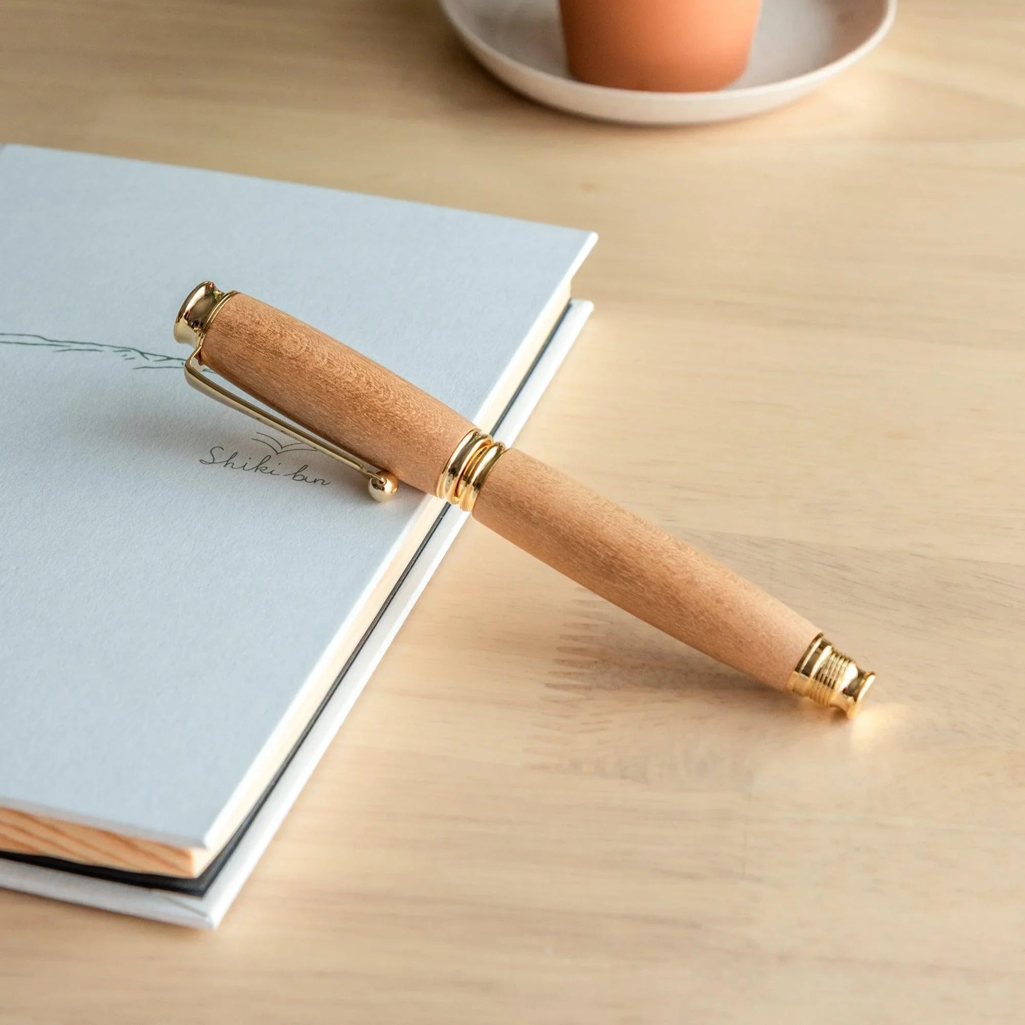 Natural wood Japanese fountain pen body uncapped on a light blue notebook on a wood desk, terracotta cup in background