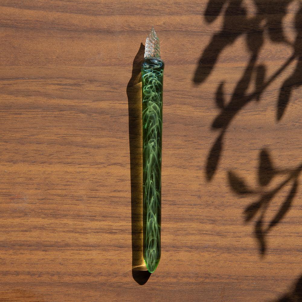 Green smoky glass dip pen standing upright on wooden surface with leaf shadow cast on white backdrop