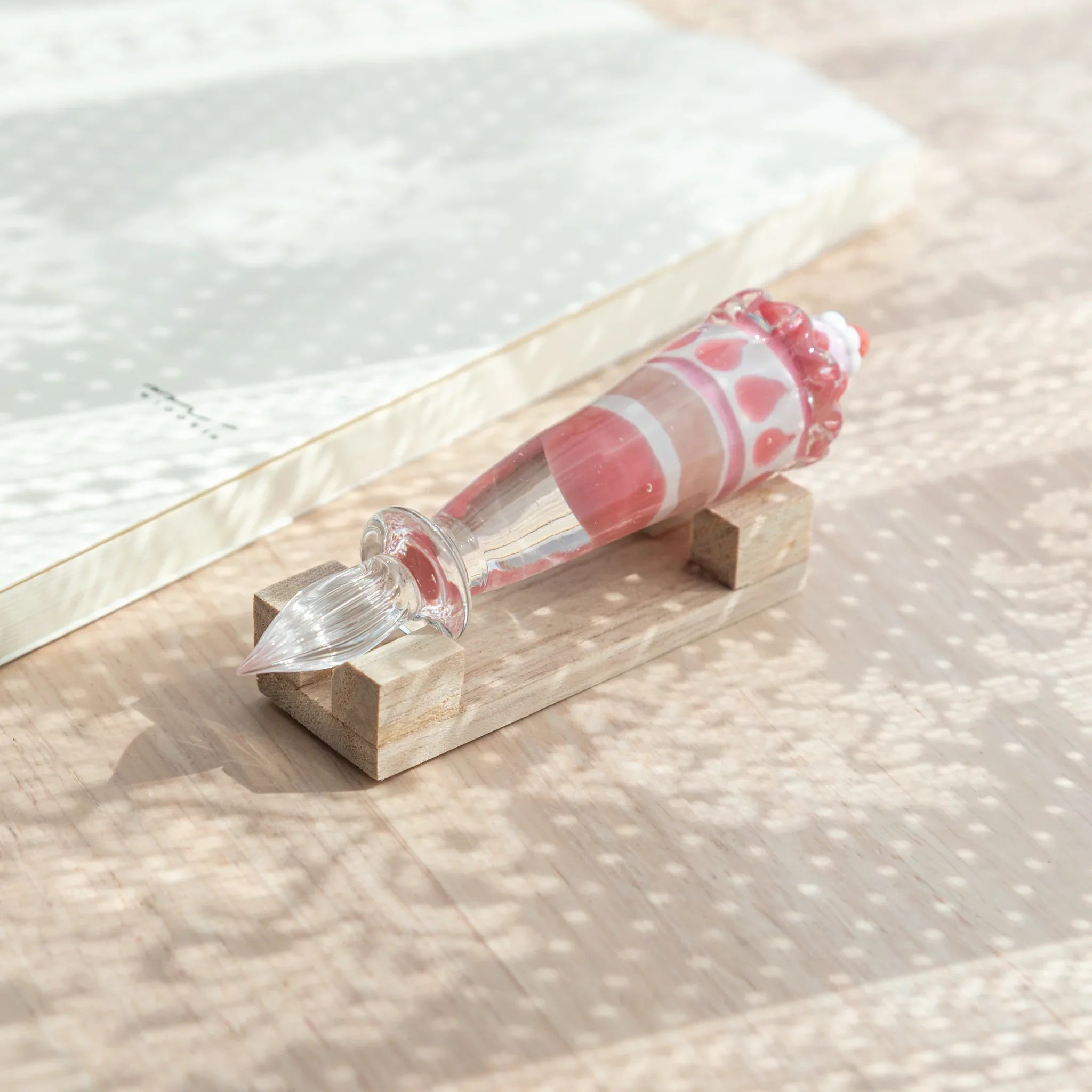 Japanese glass dip pen with strawberry parfait glass grip resting on natural wood pen rest, with open notebook in background