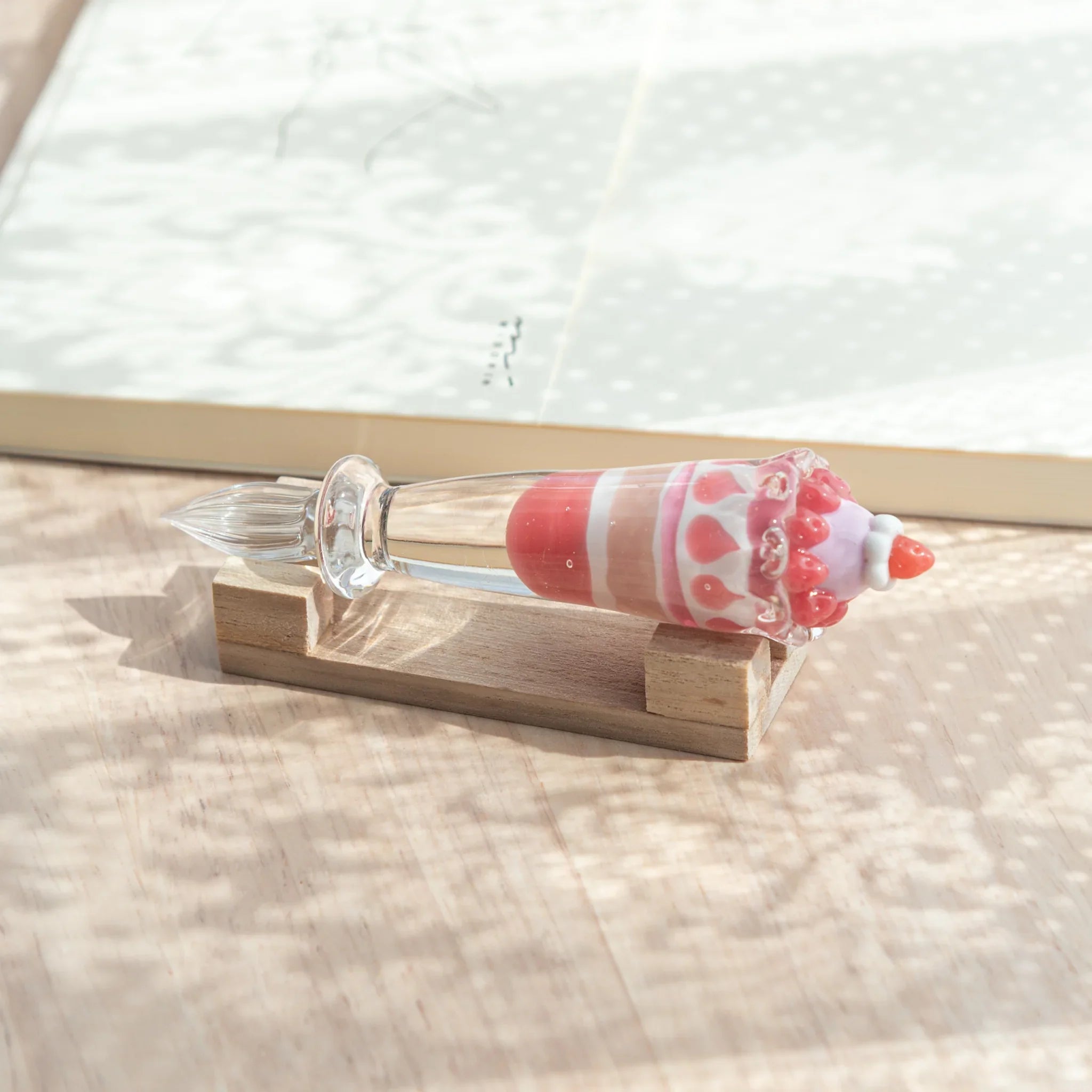 Japanese glass dip pen with strawberry parfait glass grip on natural wood pen rest, showing red and white body and clear nib