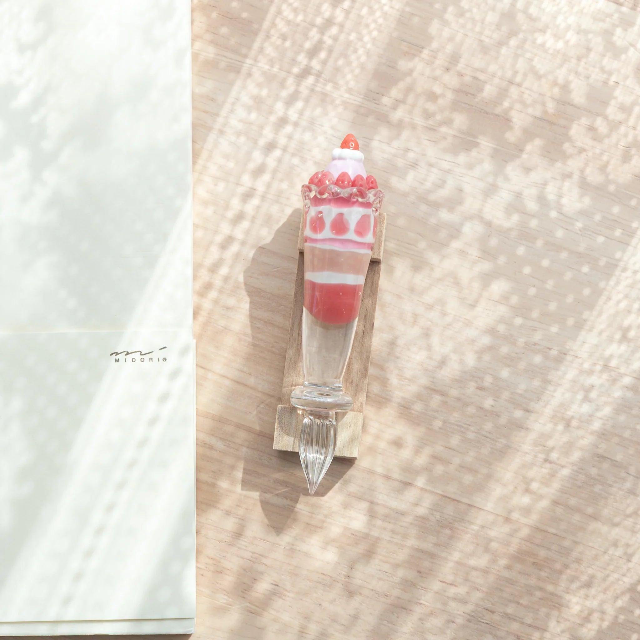 Overhead view of Japanese glass dip pen on wood pen rest with parfait grip, beside Midori notebook on light wood surface