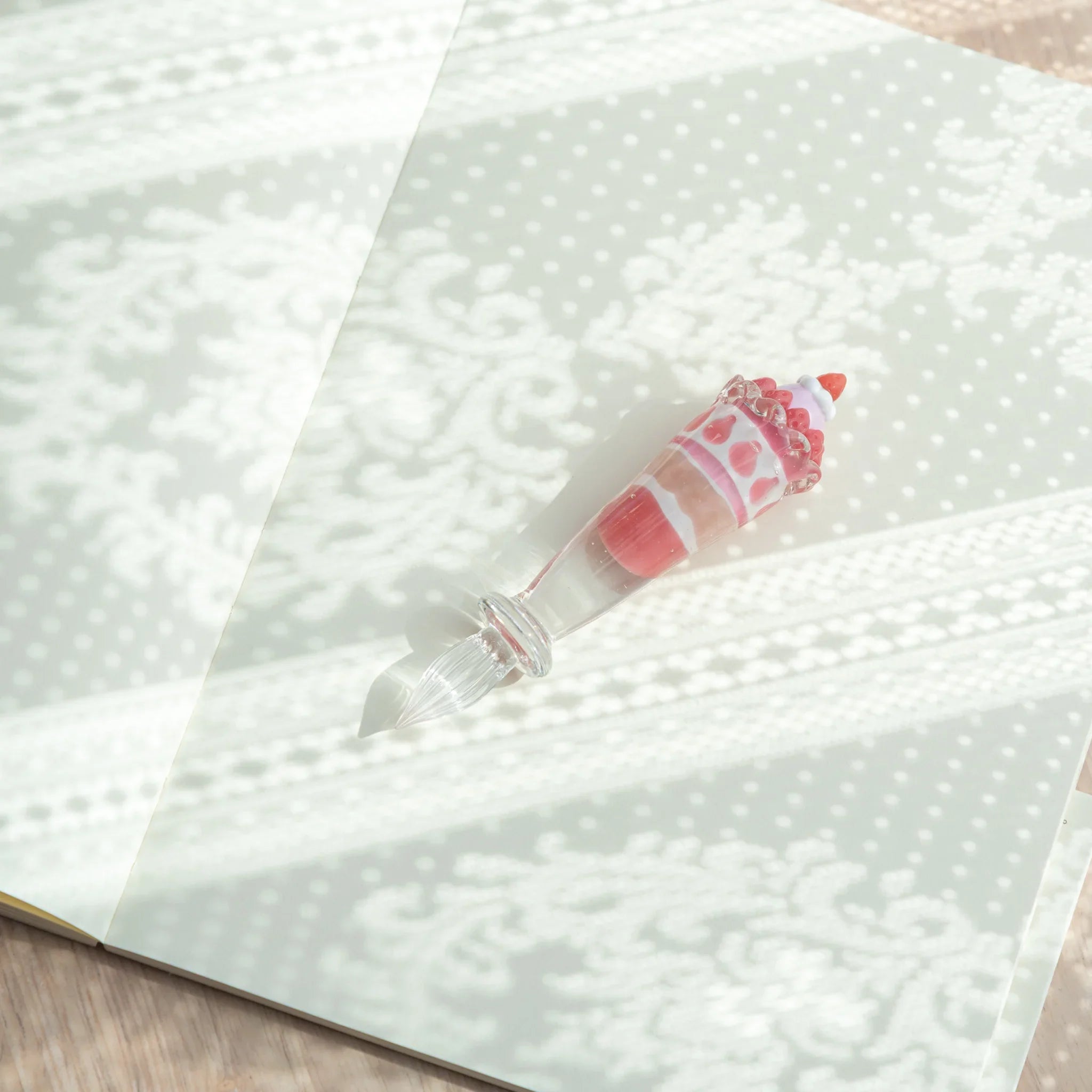 Japanese glass dip pen with strawberry parfait glass grip lying diagonally on open notebook pages with dappled light pattern